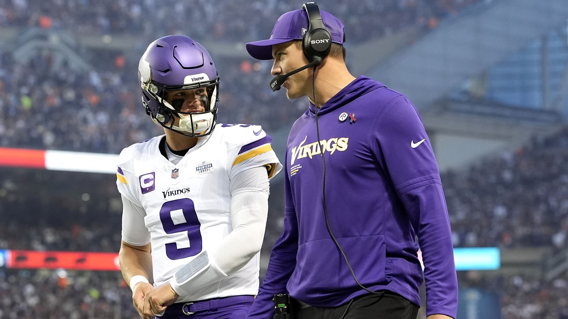 Two Vikings on the sideline: a player in a white jersey with a purple helmet talks to a coach in a purple hoodie and headset, with a blurred stadium crowd in the background.
