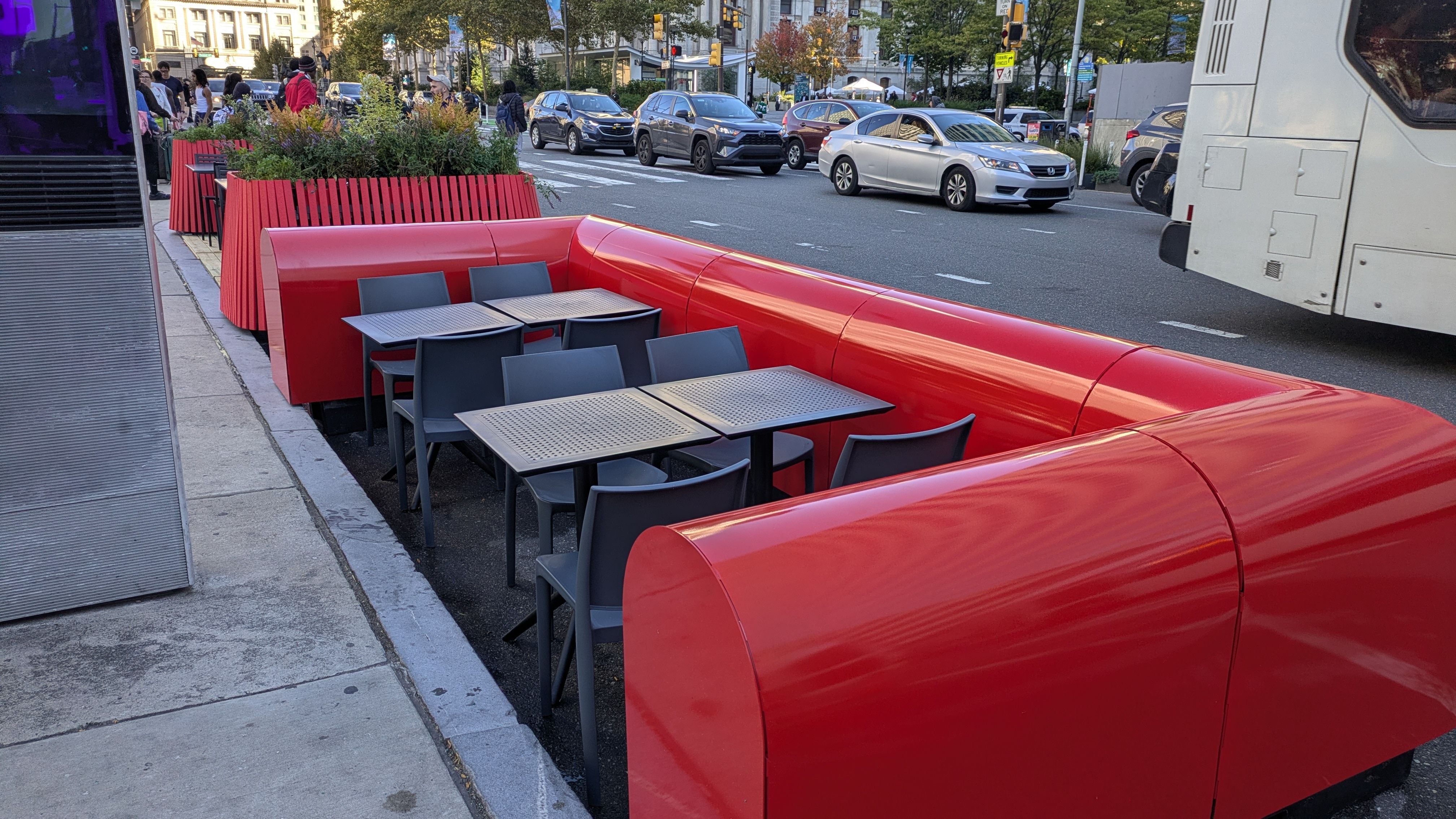 Outdoor cafe with bright red curved benches and gray tables along a city sidewalk; planters with greenery, pedestrians, and traffic, including a white bus on a busy street.
