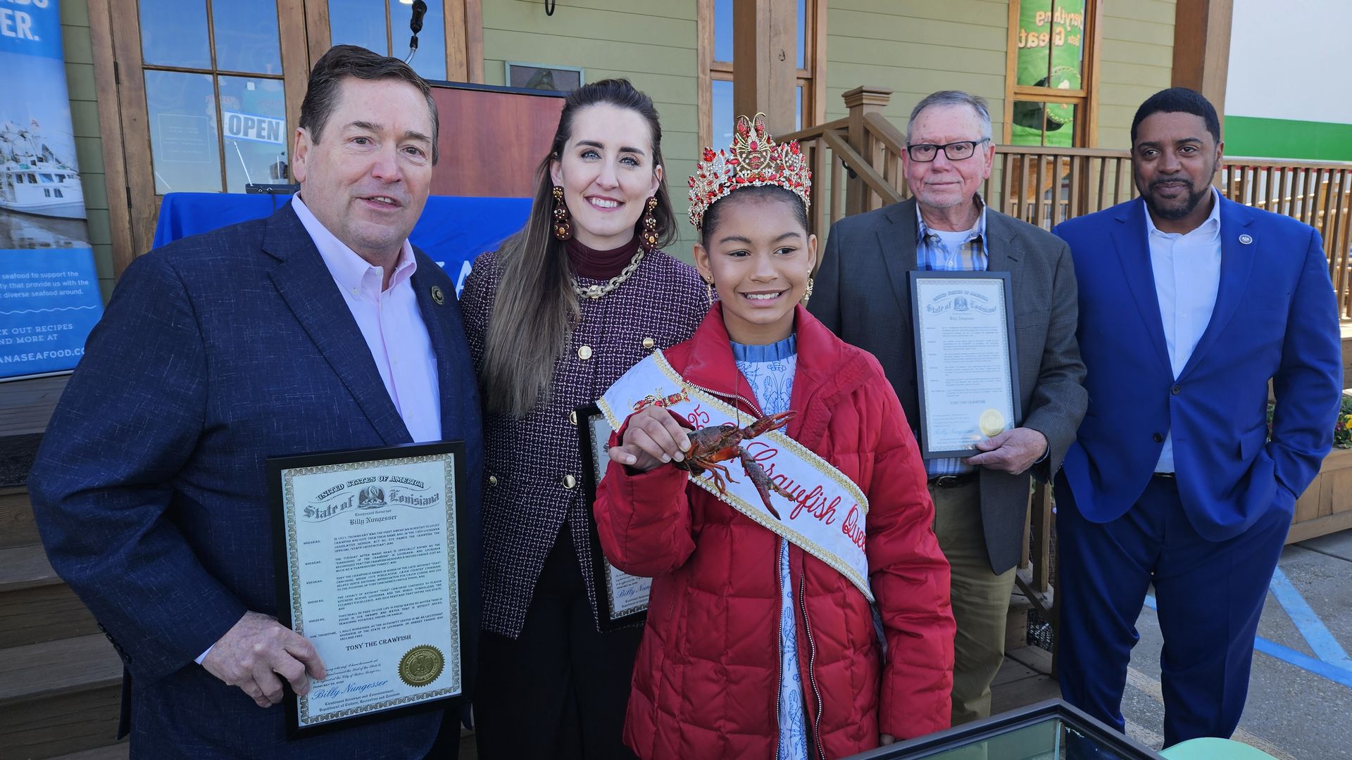 Five people stand in front of a wooden building. A girl in a red jacket and crown, wearing a sash reading "Crawfish Queen," holds a crawfish. Two men hold framed certificates.