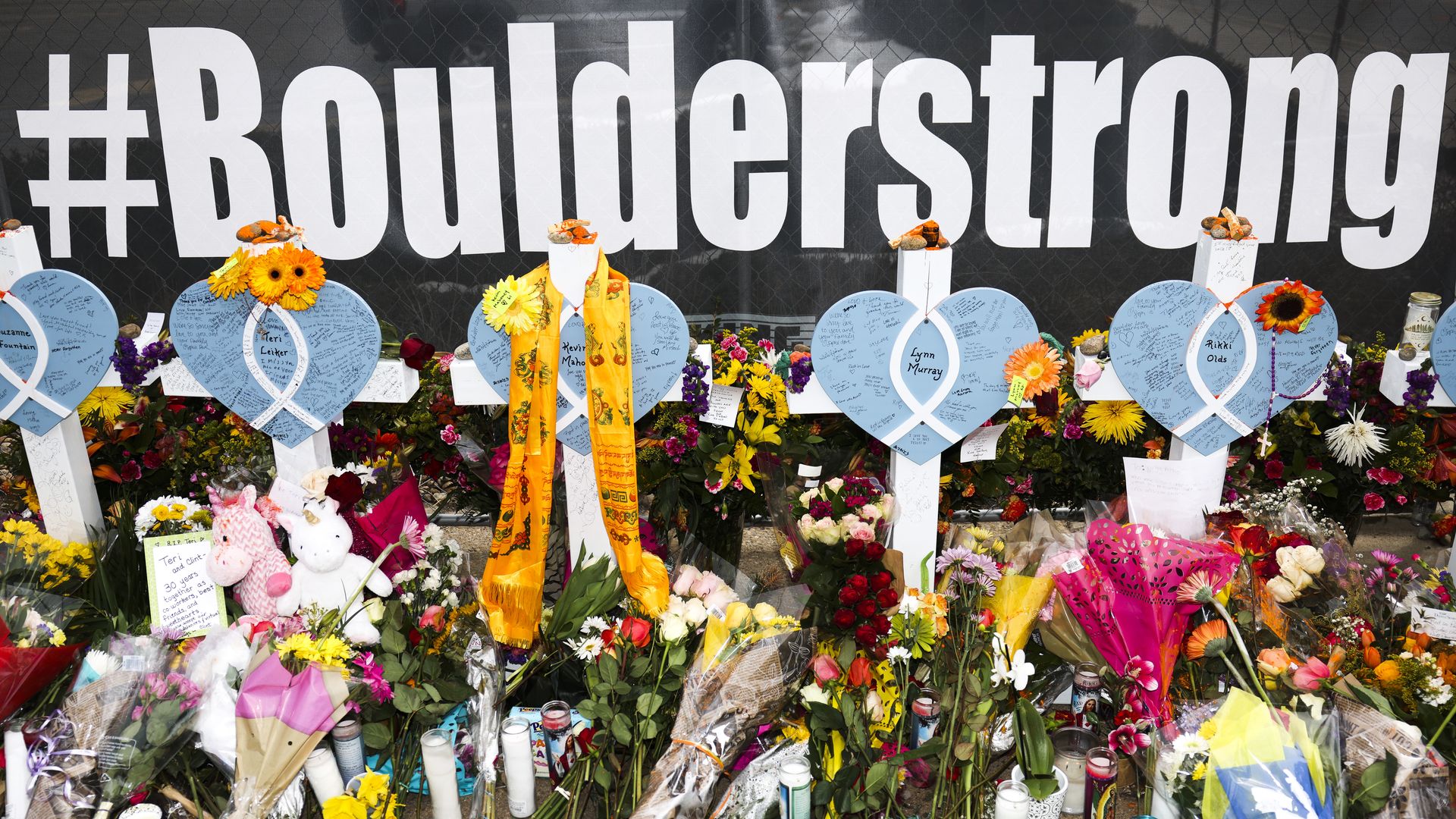 A memorial scene under a black banner with "#Bounderstrong" in large white letters. White crosses hold blue heart cards among colorful bouquets, notes, ribbons, plush toys, and candles.