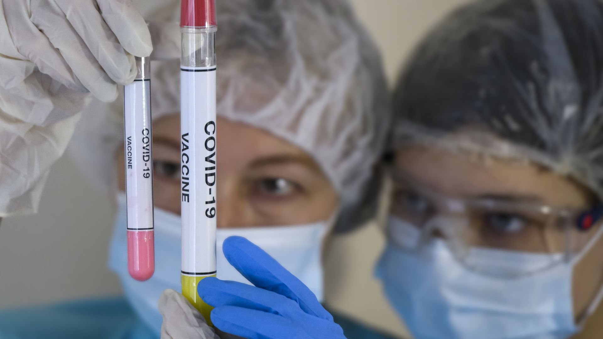 Picture of two health professionals wearing masks and other protective equipment while holding test tubes with samples of the coronavirus vaccine