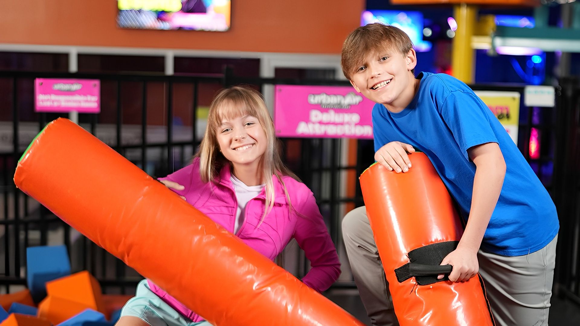 Two smiling kids at an indoor trampoline park, one in a pink jacket sitting on an orange edge, the other in a blue shirt kneeling, both holding large orange padded sticks above blue and orange foam cubes.