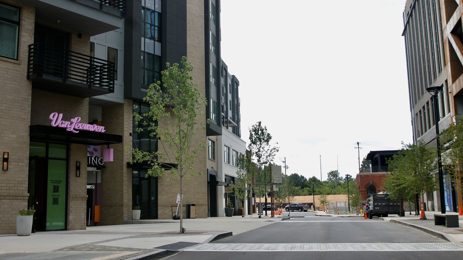 Urban street view with modern buildings, small trees along sidewalks, parked vehicles, and a sign reading "Van Leeuwen" on the left building in pink neon light.