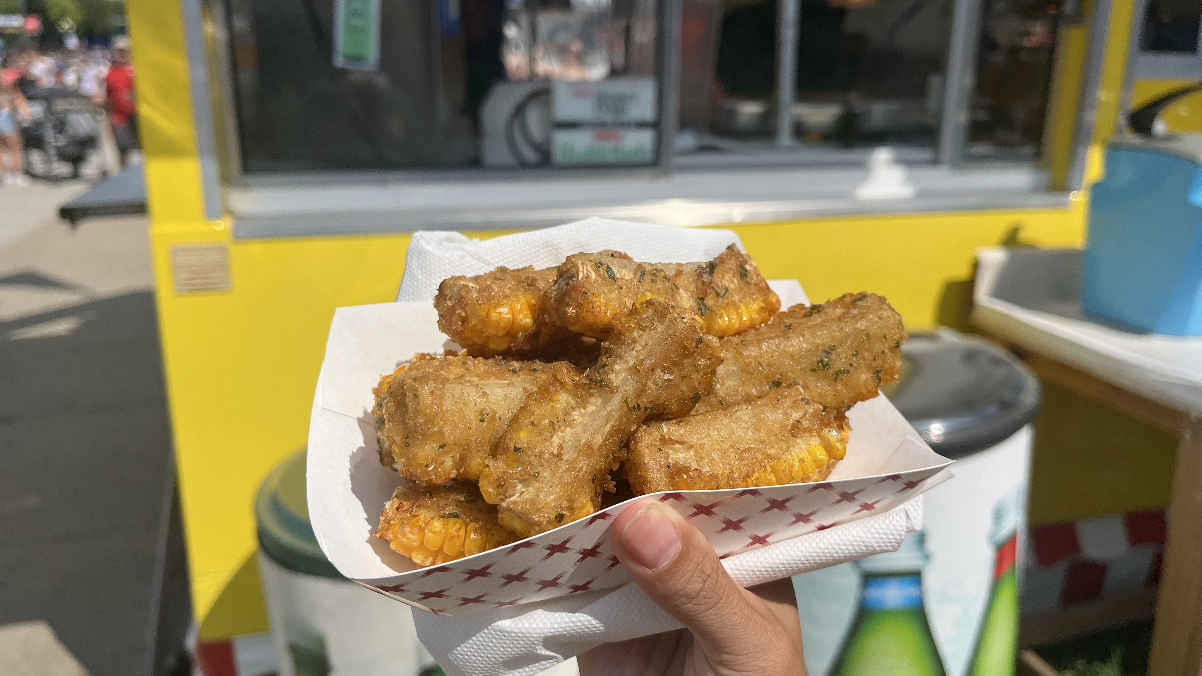 Hand holding a tray of fried corn on the cob pieces coated with herbs and batter, in front of a yellow food truck.