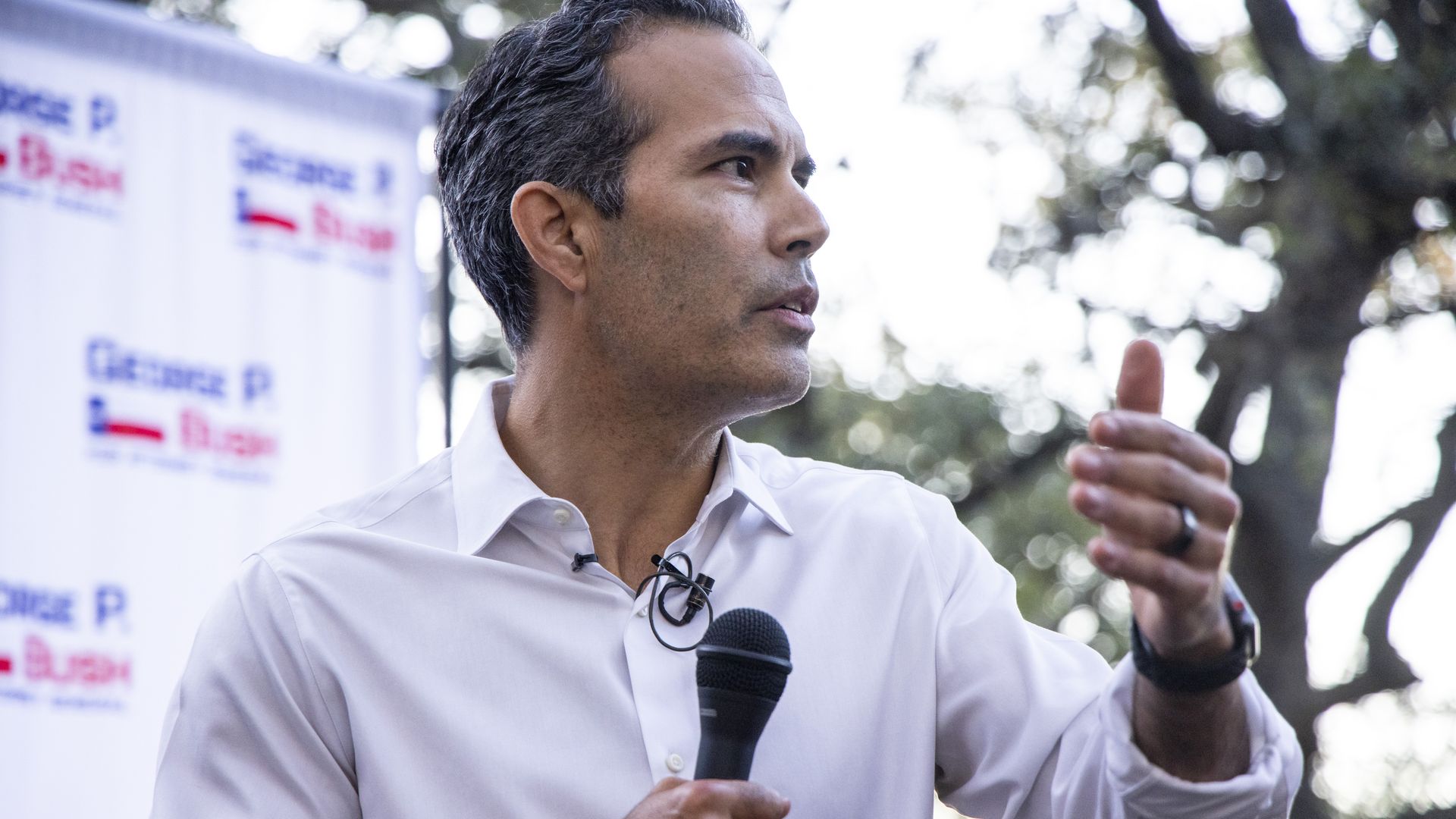 George P. Bush speaks at an outdoor campagin event while holding a black microphone in one hand and gesturing with the other