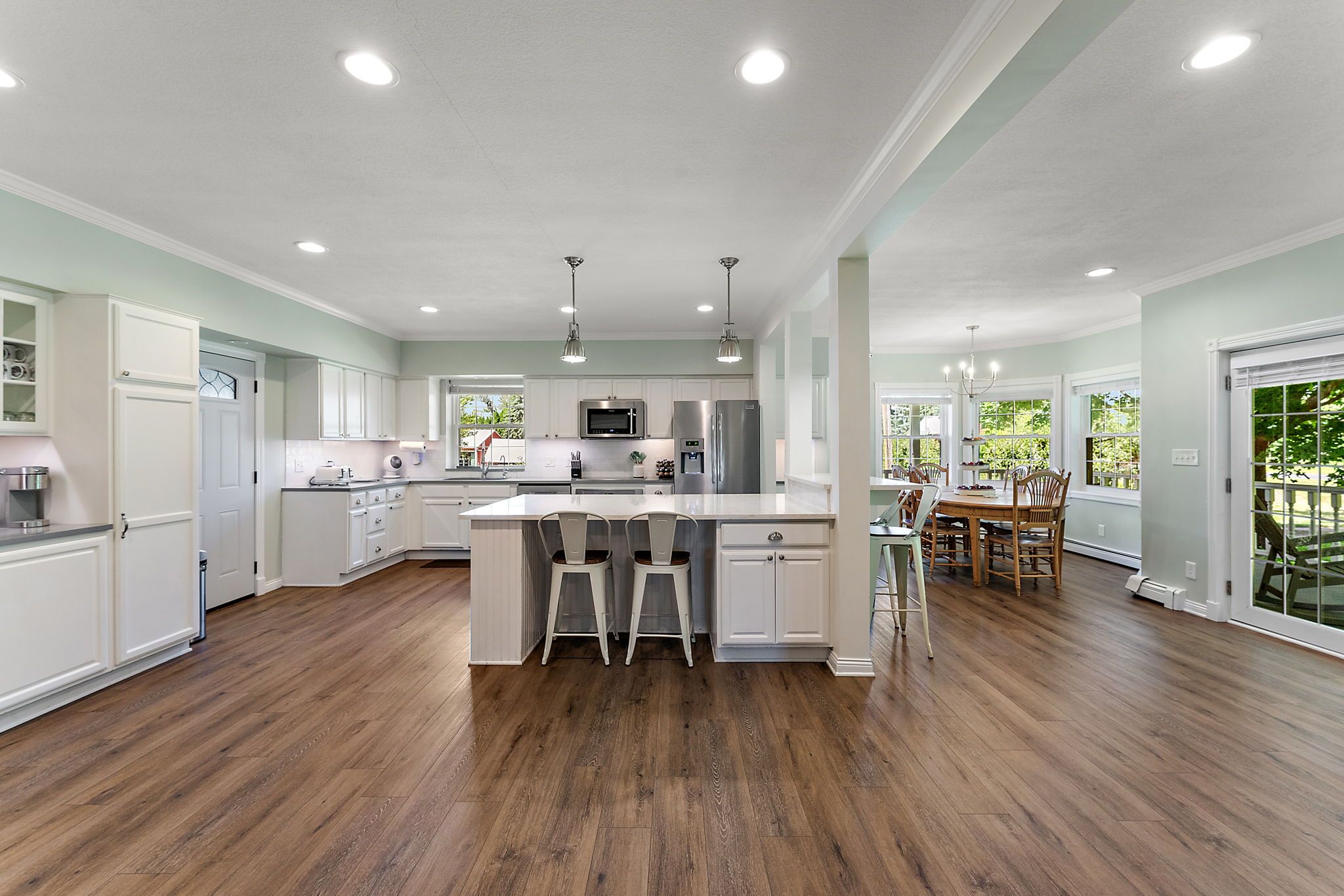 Spacious kitchen and dining area with white cabinets, two metal stools at island, wooden dining table, green walls, large windows, and wood flooring under bright ceiling lights.