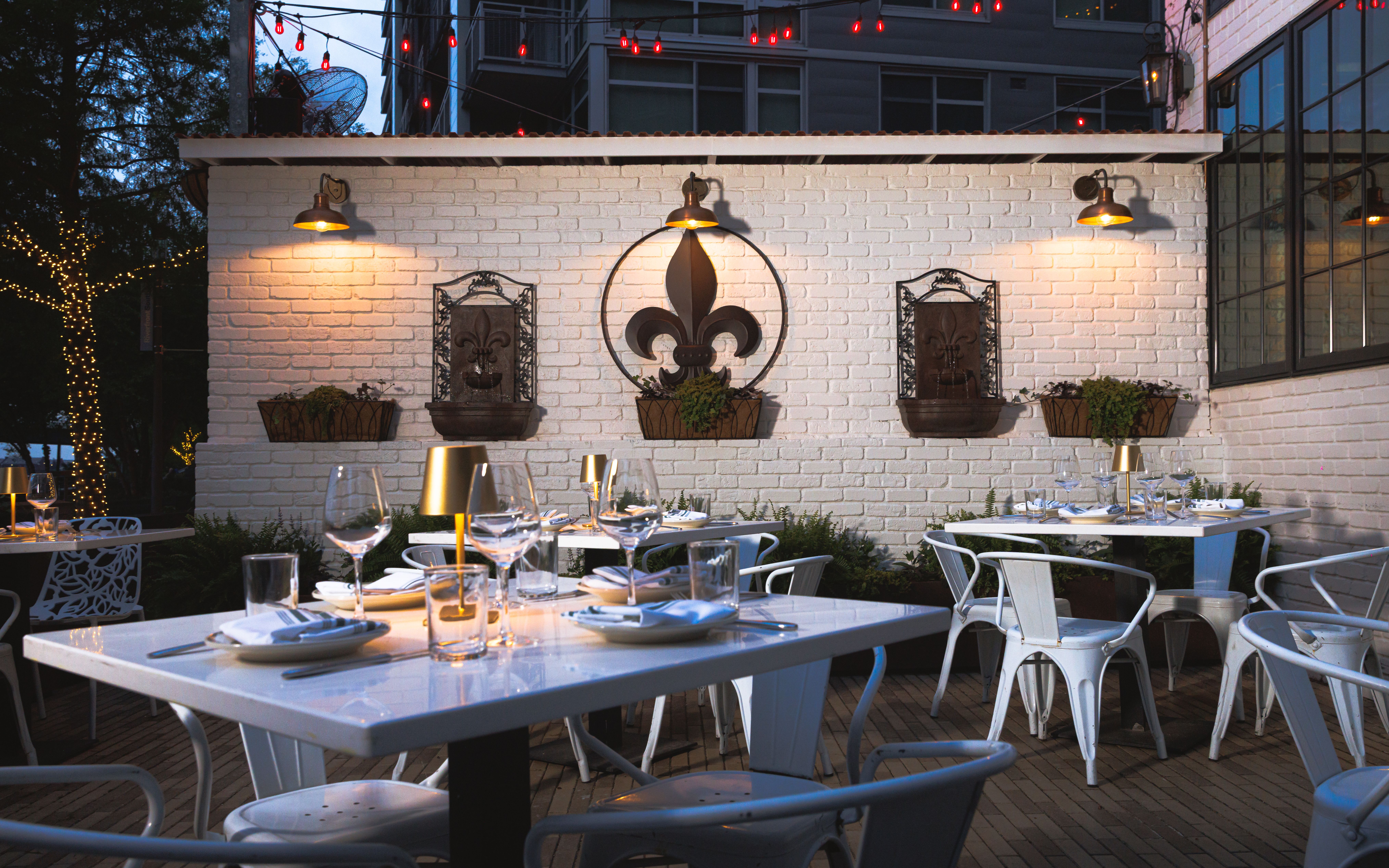 Outdoor dining area at dusk with white brick wall, fleur-de-lis emblem inside a circle, two ornate metal wall planters, string lights, warm wall lamps, and tables set with glassware and plates.