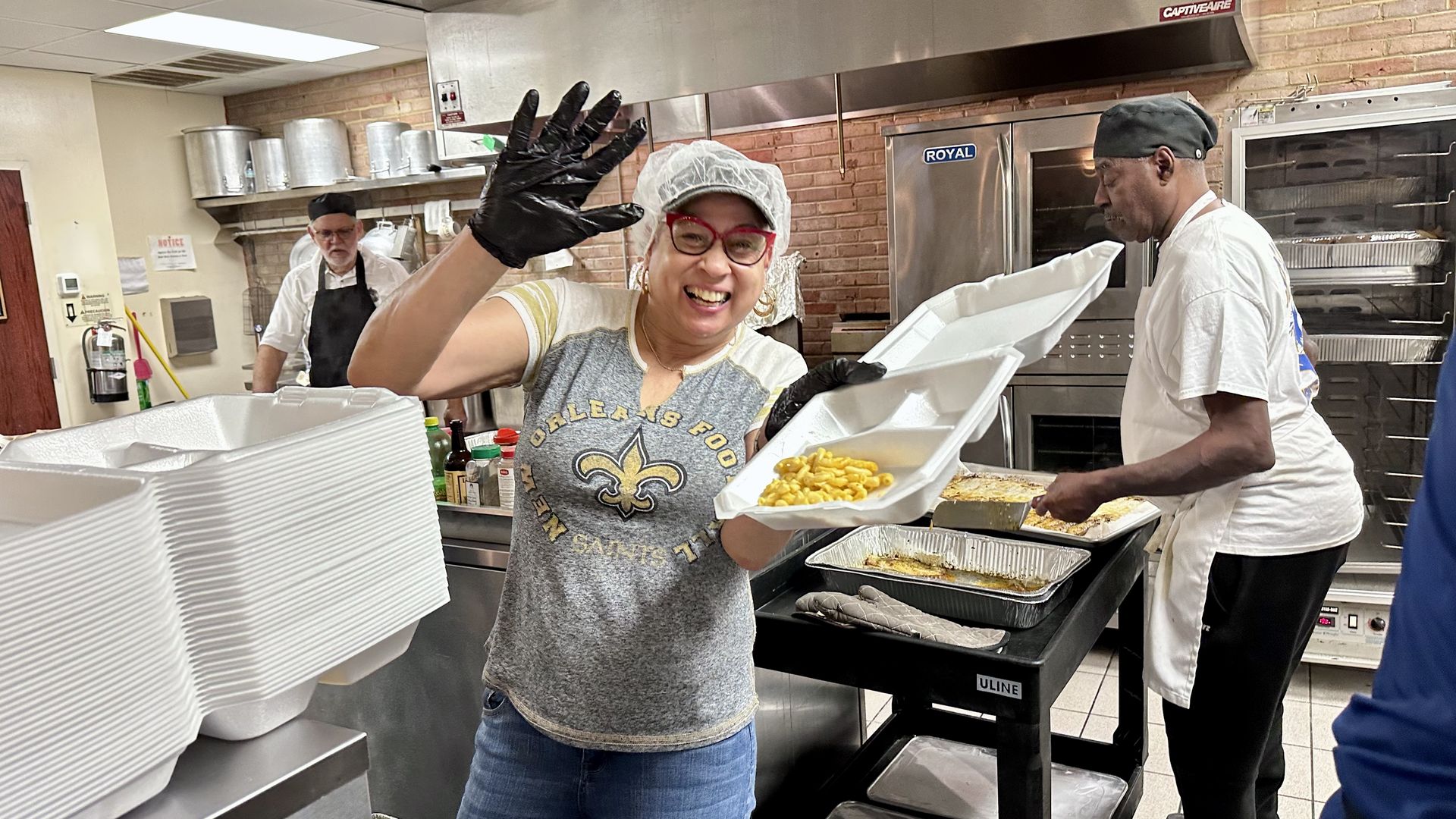 Photo shows a woman smiling and waving with a box of fried fish and mac and cheese.