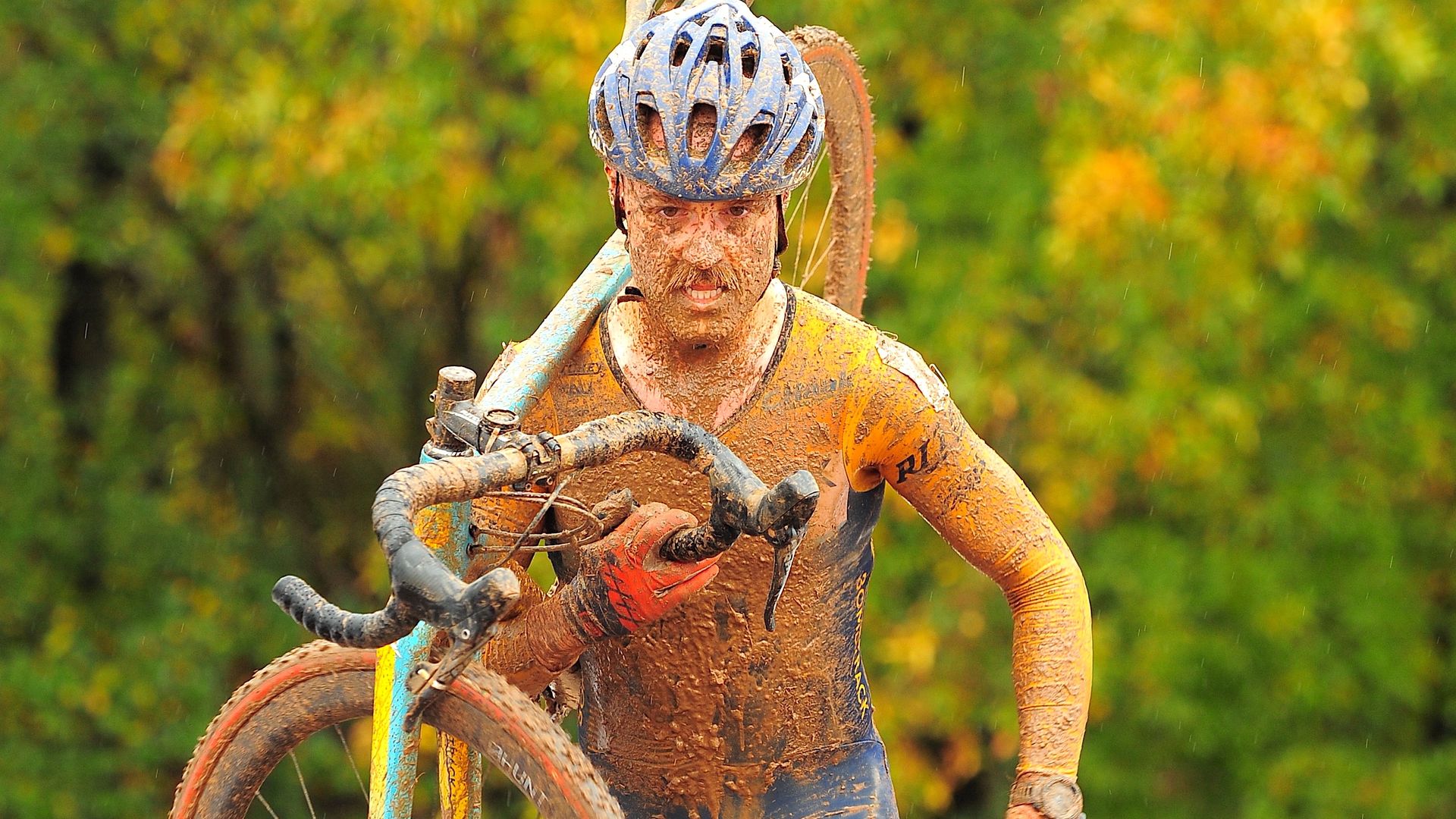 A man covered in mud, wearing cycling gear and a helmet, carries a bike on his shoulder in the rain.