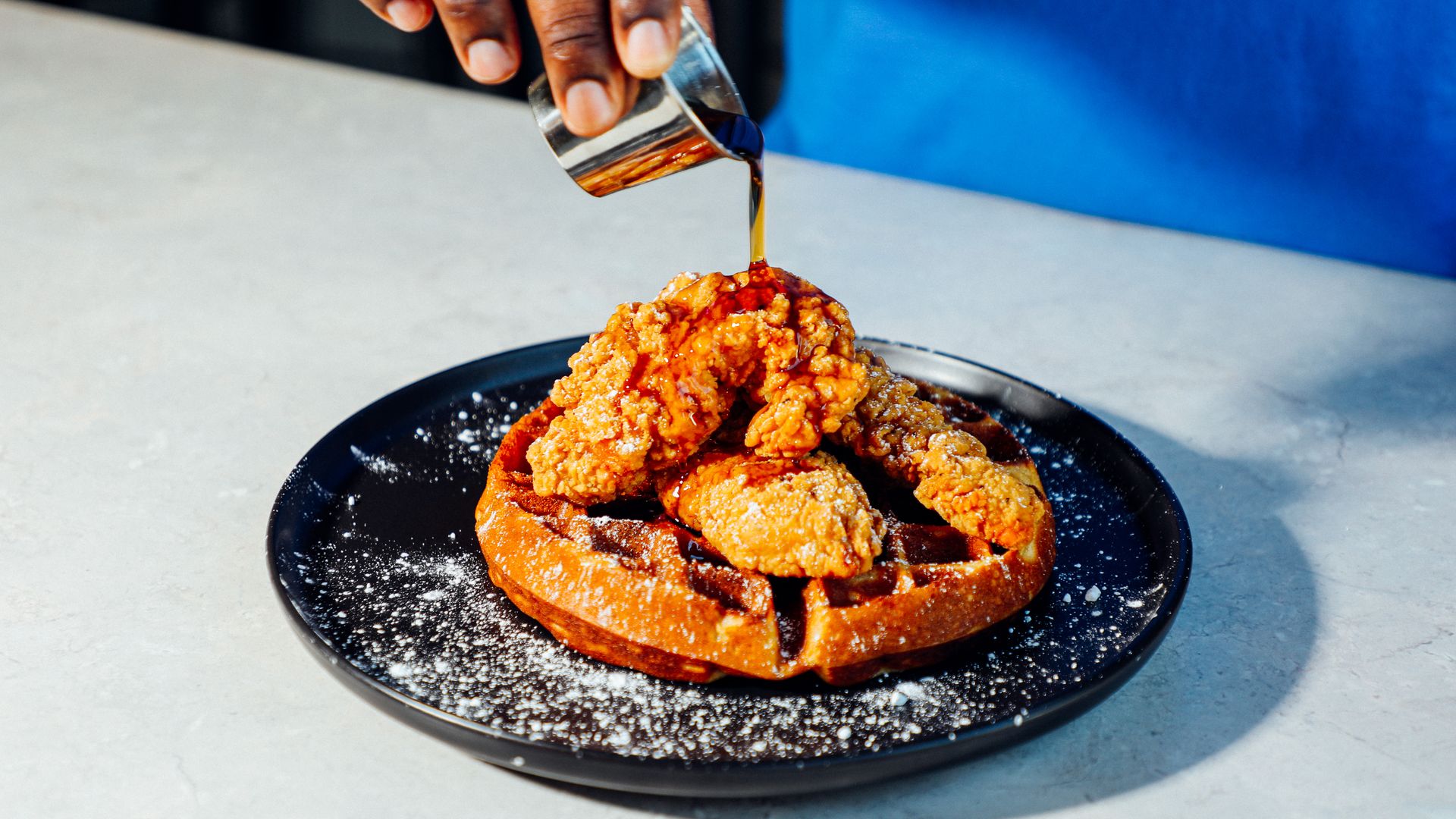 A person pours syrup on chicken and waffles on a black plate with a white counter underneath 