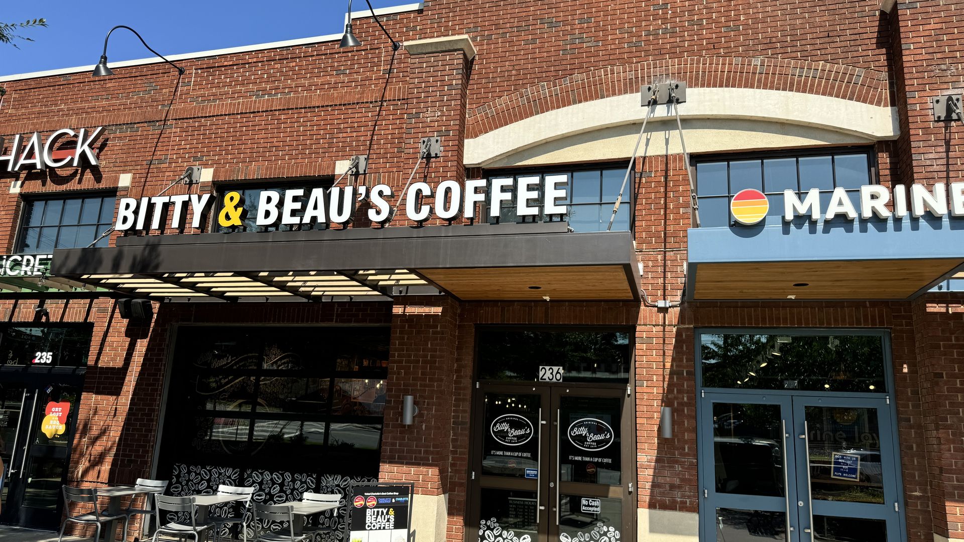 Exterior of Bitty & Beau's Coffee in a red brick building with black awning, outdoor seating with metal chairs, and bright blue sky overhead.