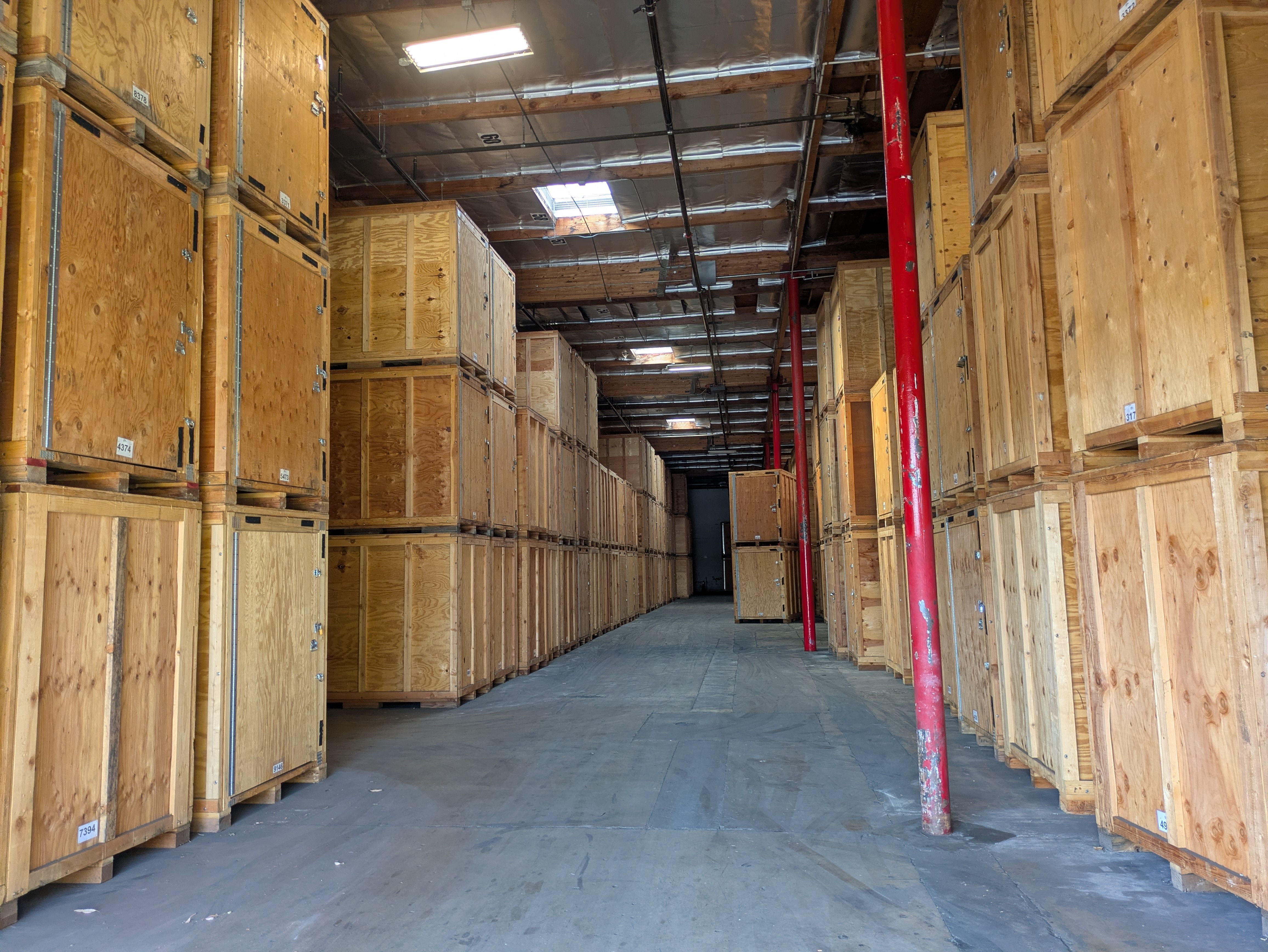 Warehouse interior with tall stacks of wooden storage crates lined up on both sides and red metal support beams, under a ceiling with exposed pipes and skylights.