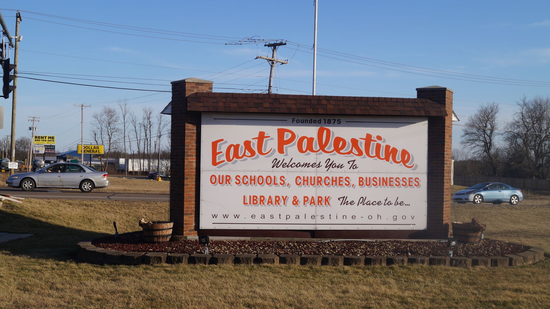 Welcome sign for East Palestine, Ohio, in orange and black letters on white, surrounded by brick pillars, promoting schools, churches, businesses, library, and park under blue sky.