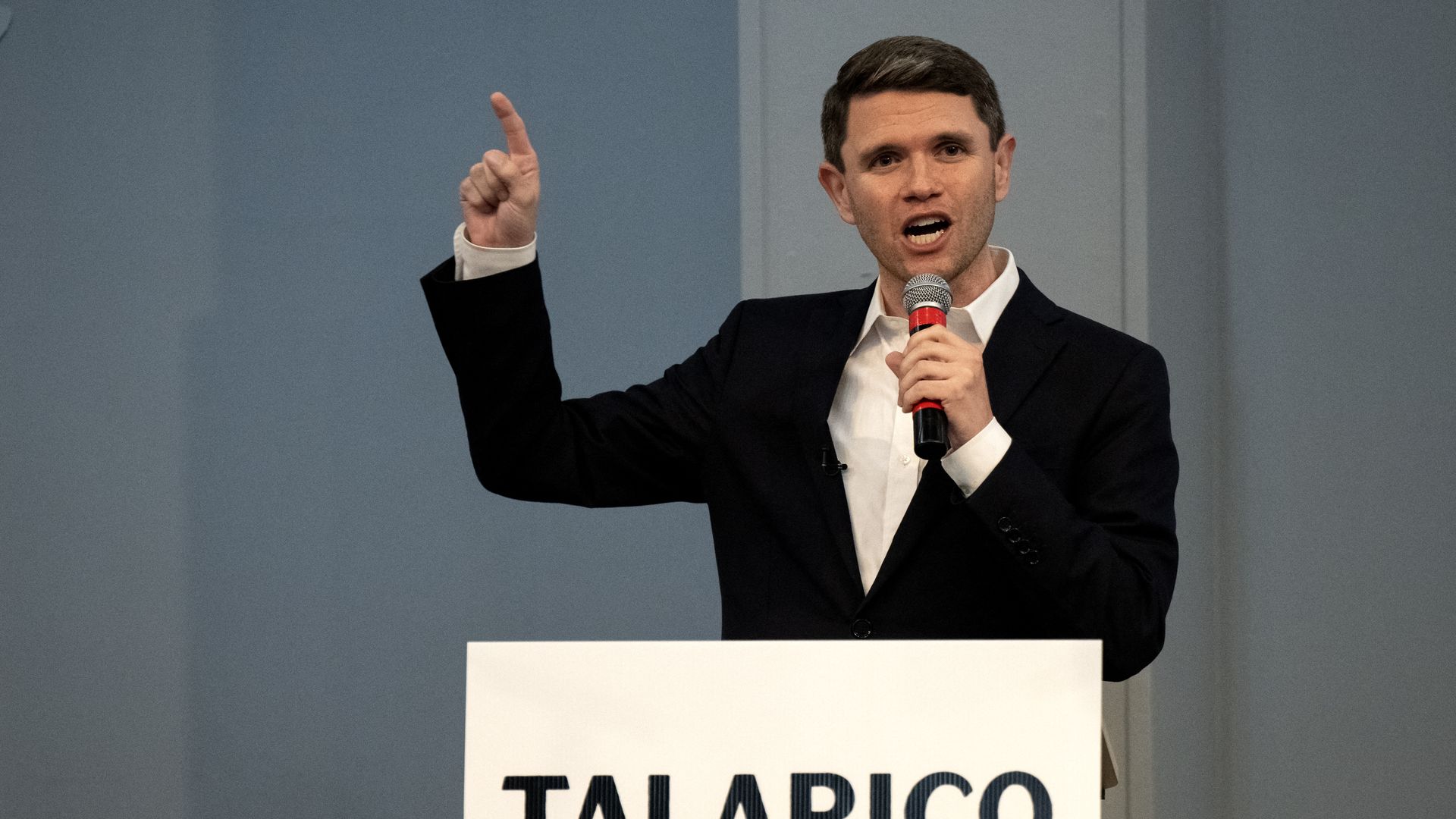A man in a black suit and white shirt speaks at a podium, one hand raised with index finger pointing up, the other holding a microphone. The podium sign reads "TALARICO" and "FOR TEXAS".