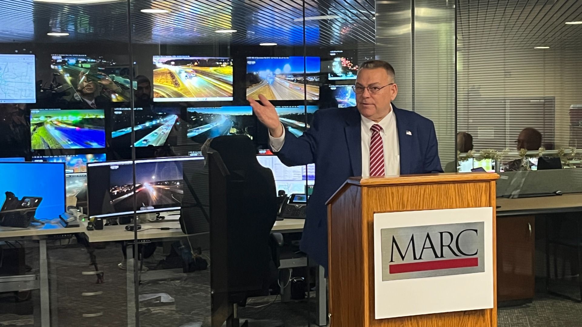 Lee White stands in blue suit and red striped tie speaking at a wooden podium with "MARC" logo, multiple surveillance screens showing highway traffic behind glass in a control room.