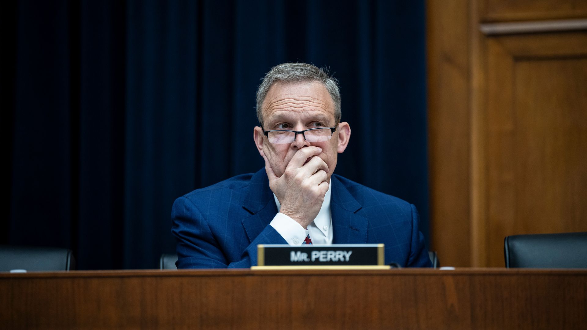 Rep. Scott Perry, wearing a blue suit and glasses with his face in his hand, sitting at a wooden dais with a plaque reading "Mr. Perry," in front of a blue curtain and a wooden wall.