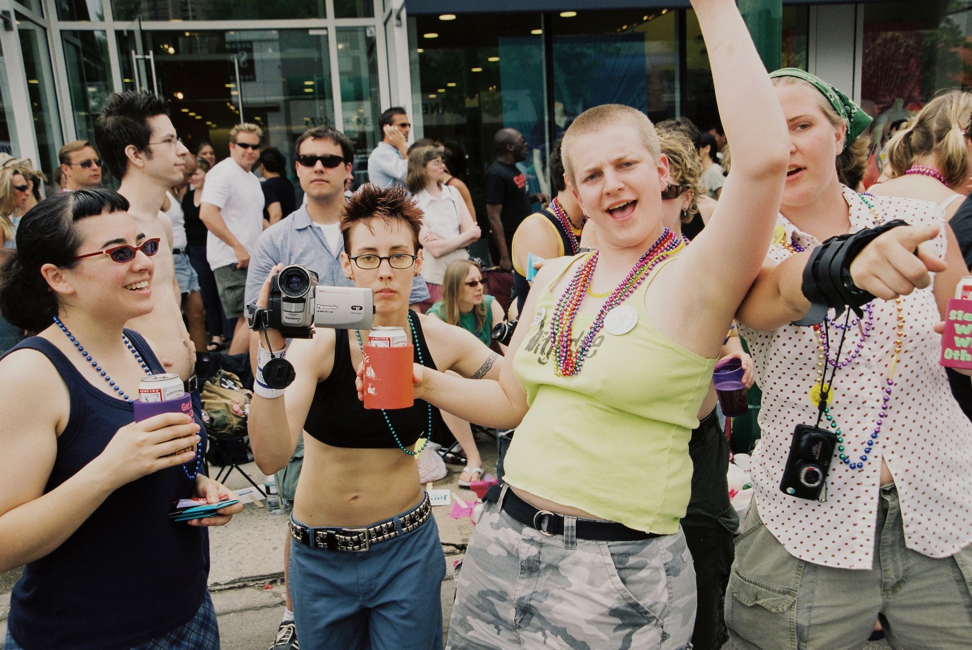 A dark-haired person in sunglasses and a tank top smiles at a friend, a  person with just a sports bra and buzz cut holds a camcorder at the photographer, and a person with a blond buzz cut, tank top and beads has one arm up and mouth open, and a drink in the other hand. 