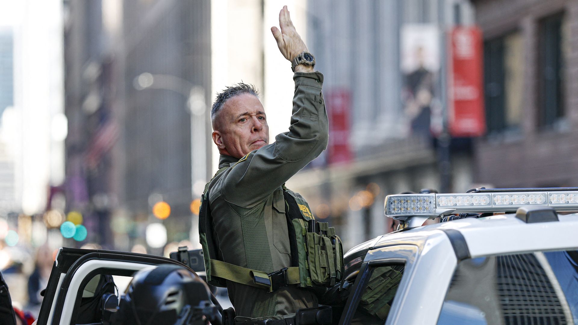 Gregory Bovino getting into a car and turning his head toward the camera with his right arm raised.