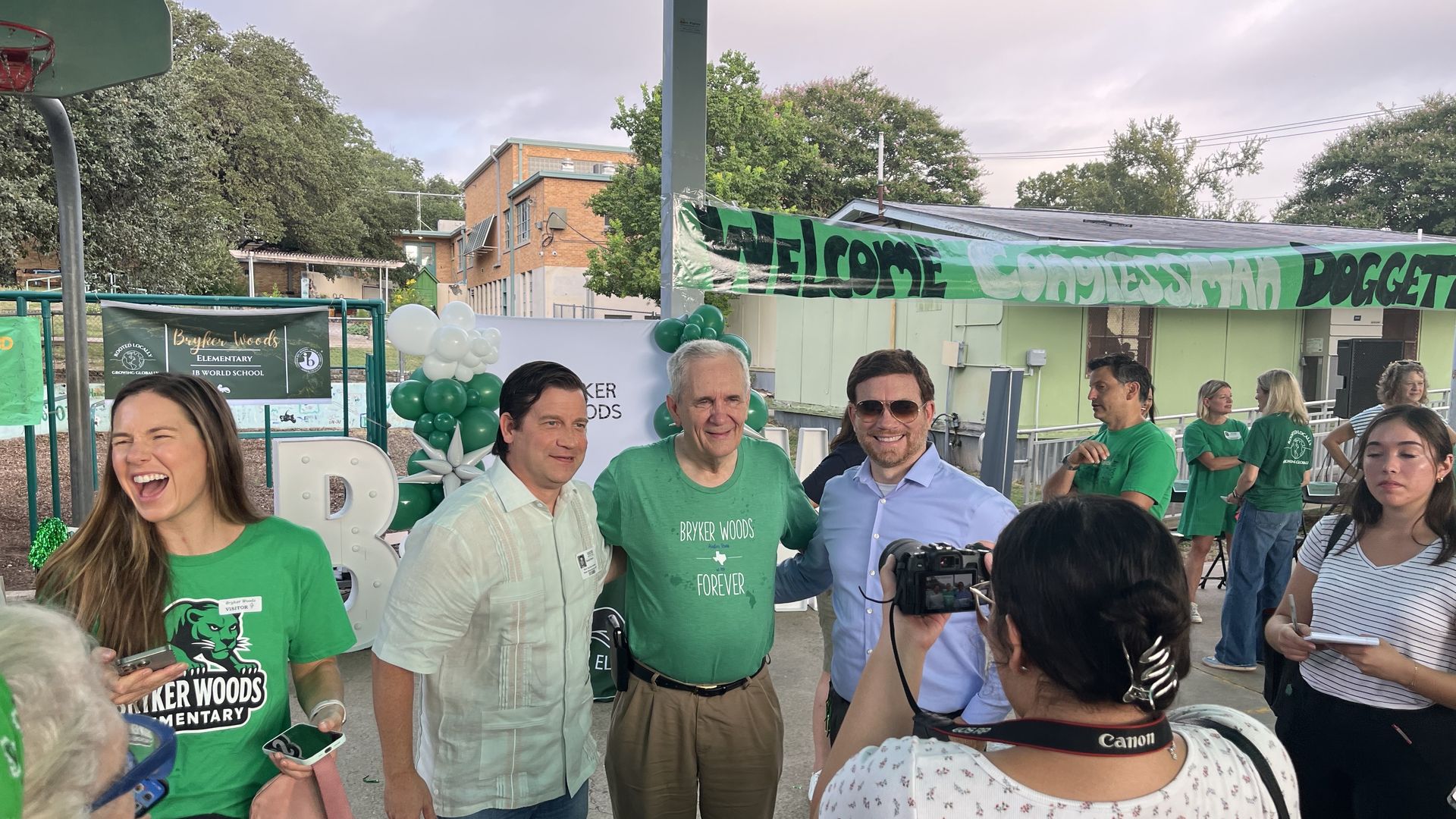A group of smiling adults including three men and one woman posing for a photo at Bryker Woods Elementary event with green and white balloons, banners, and attendees in green shirts.