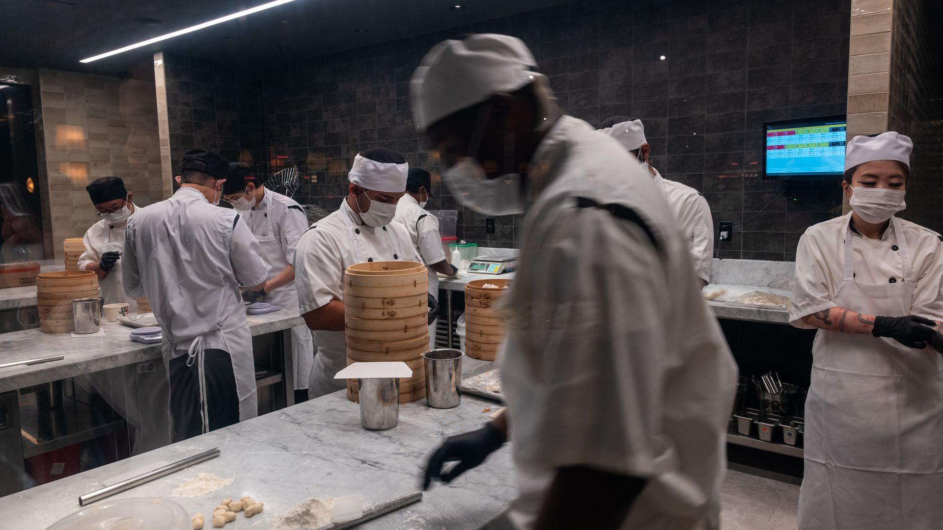 Busy professional kitchen with chefs in white uniforms and masks at a marble counter. Stacked steamers and dough on the counter; dark tiled walls and a digital screen in the background.