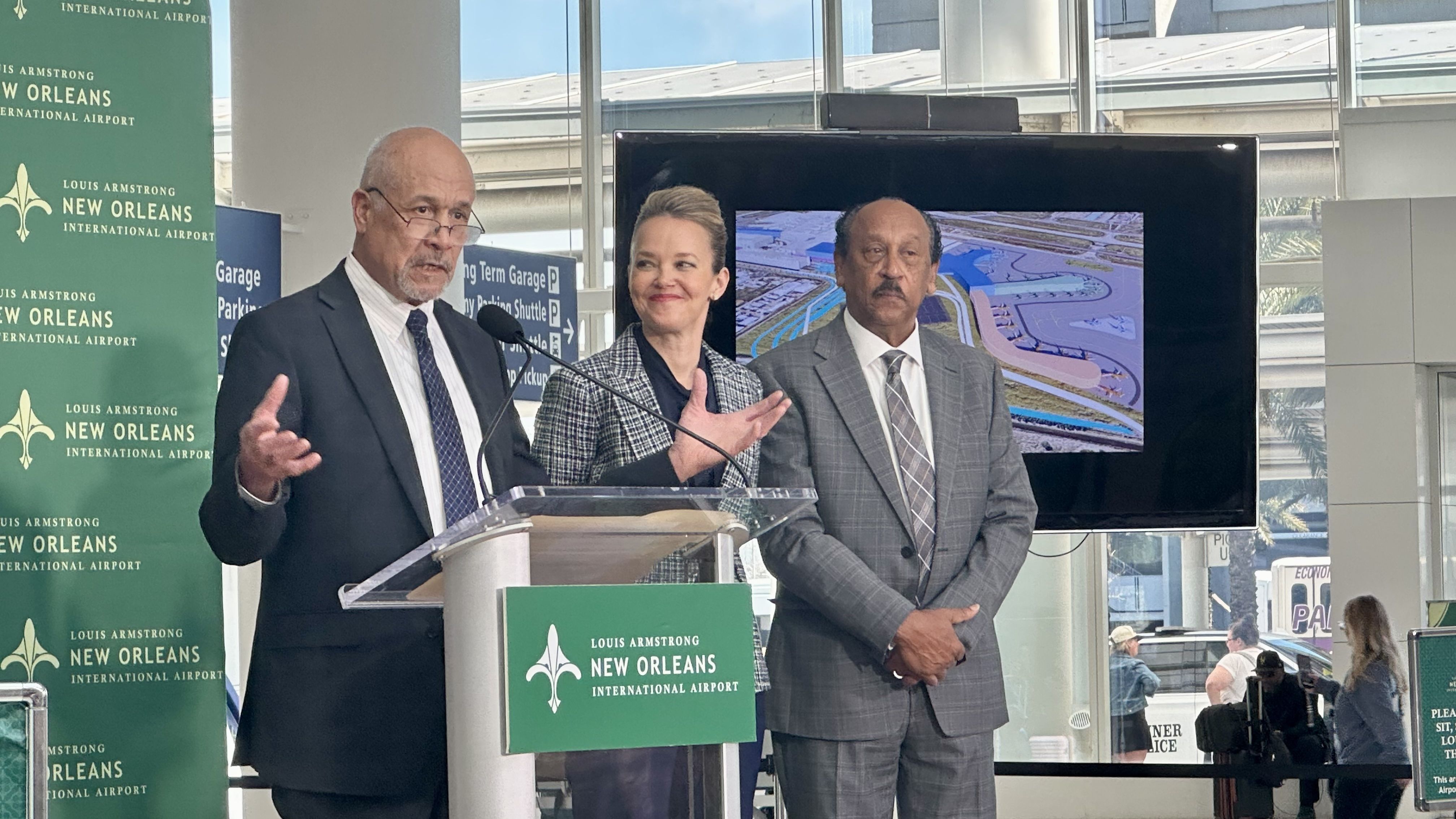 Press briefing at Louis Armstrong New Orleans International Airport. Three suited people stand at a podium: left speaker, center smiling woman in a checkered blazer, right man in gray suit; green banners and a large screen behind.