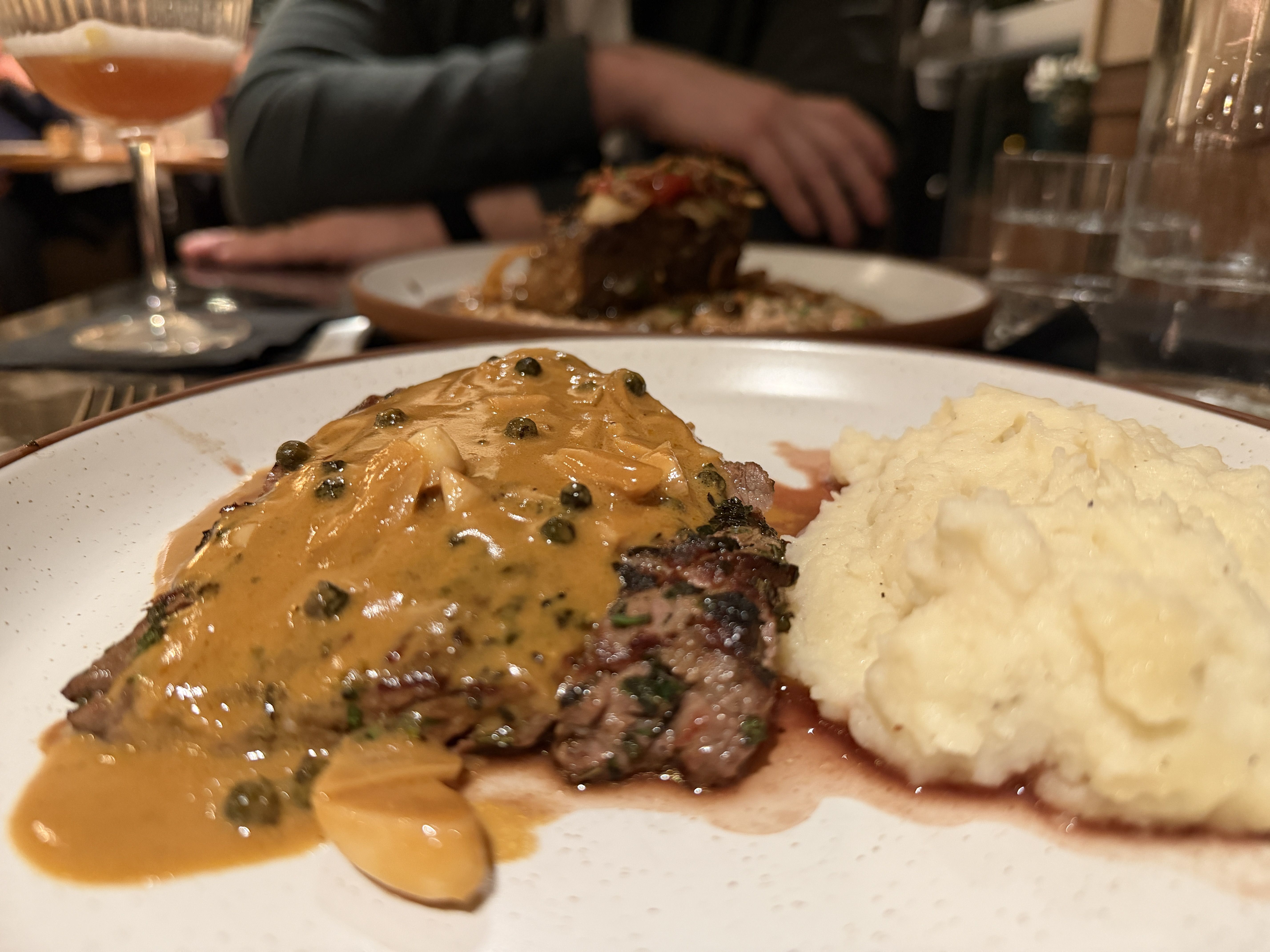 Plate with grilled steak covered in peppercorn sauce beside creamy mashed potatoes, glass of amber drink and blurred person in background at restaurant table.