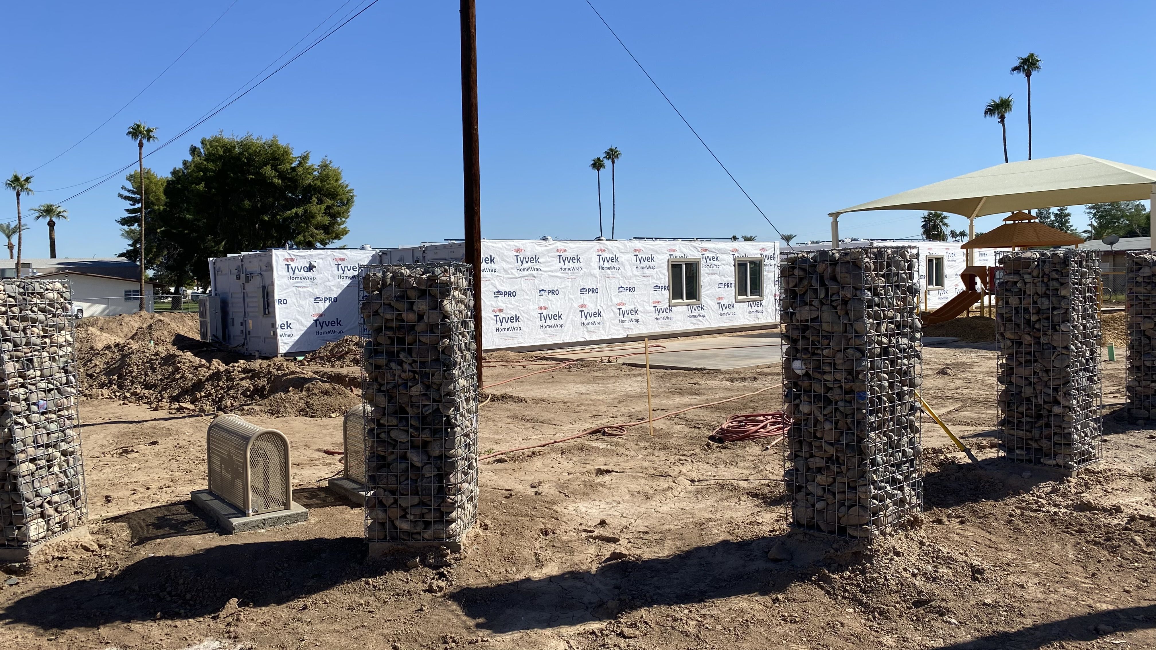 Several shipping containers being renovated at a construction site, surrounded by large metal pylons filled with rocks. 