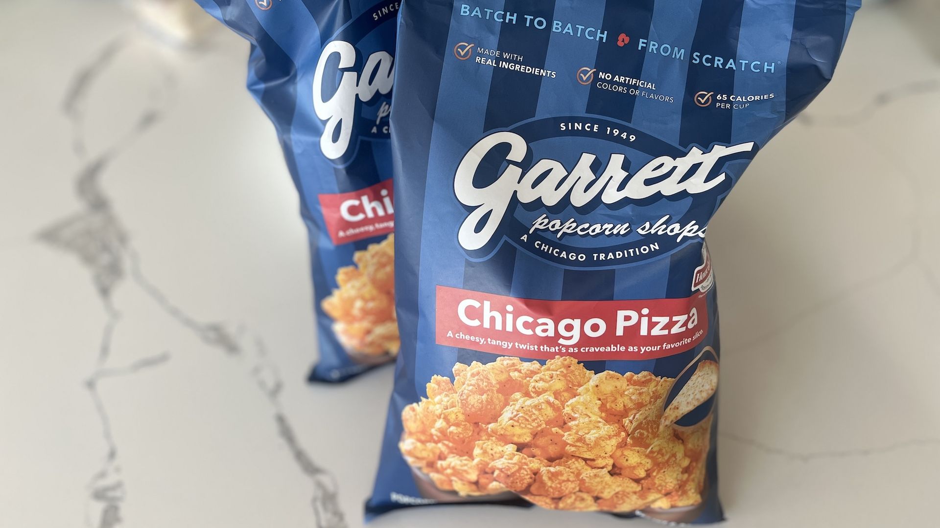 Two blue bags of Garrett popcorn, Chicago Pizza flavor, highlighting real ingredients, no artificial colors, and 65 calories per cup, on a white marble surface.