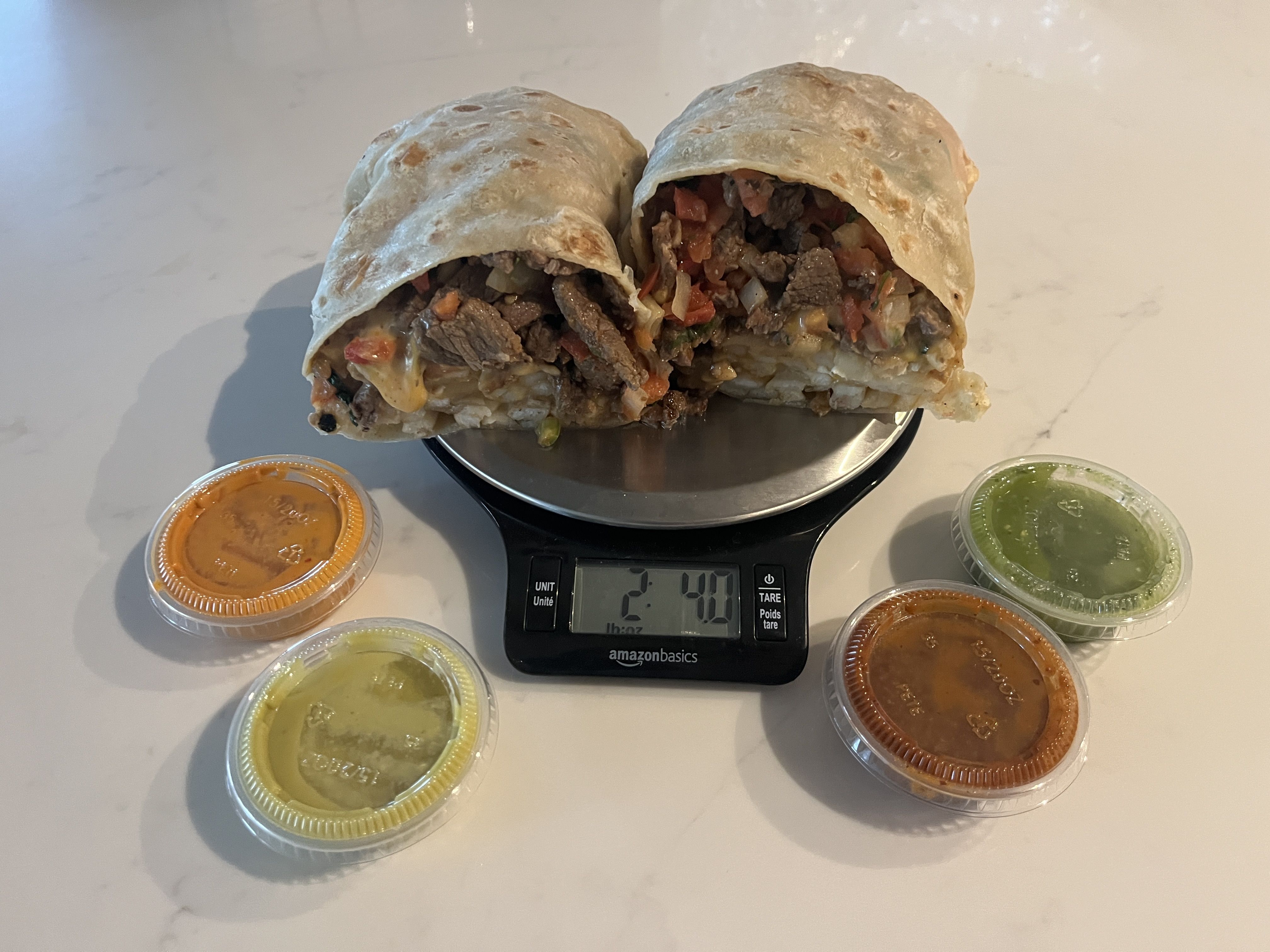Two halves of a beef burrito with melted cheese and vegetables on a digital kitchen scale reading 2lbs, 4 oz., surrounded by four small containers of orange, green, and red salsa on a white surface.