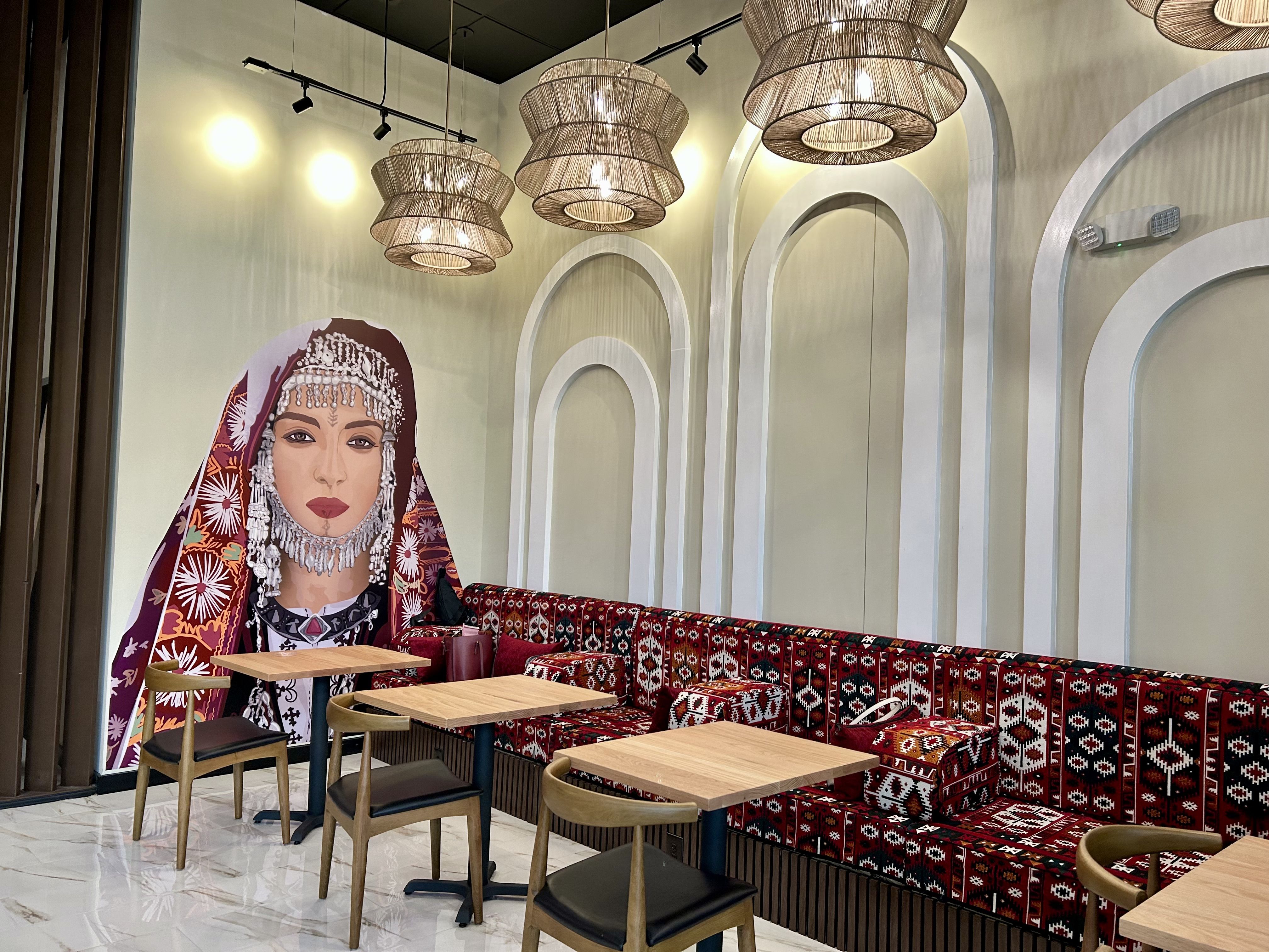 Interior of a café with wooden tables, chairs, and a long bench with red and black patterned upholstery. A wall features a large mural of a woman in traditional attire with silver jewelry.