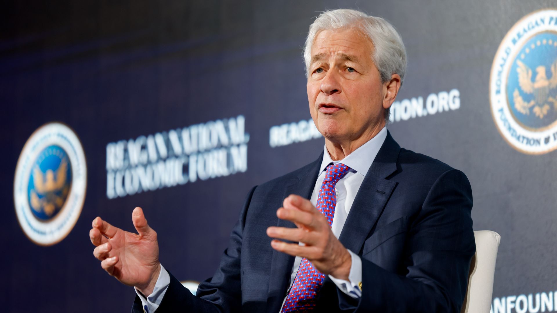 A person with white hair, wearing a dark suit and red tie, speaks on a stage while gesturing with his hands. A blue backdrop shows circular logos and the words "NATIONAL FOUNDATION".