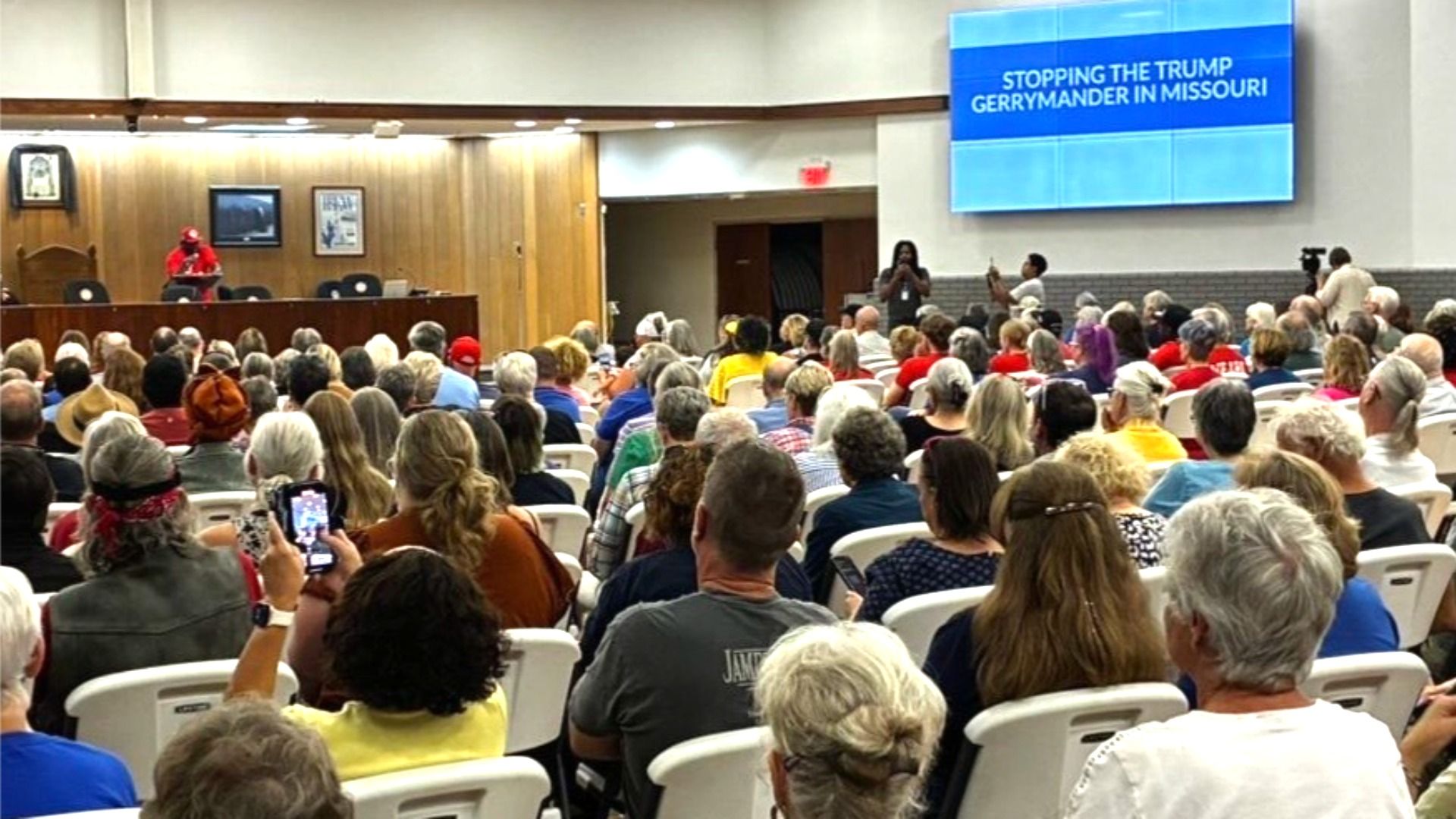 Crowd of people seated in a hall, facing a stage and a large screen displaying "Stopping the Trump Gerrymander in Missouri." A speaker in red stands at the podium.