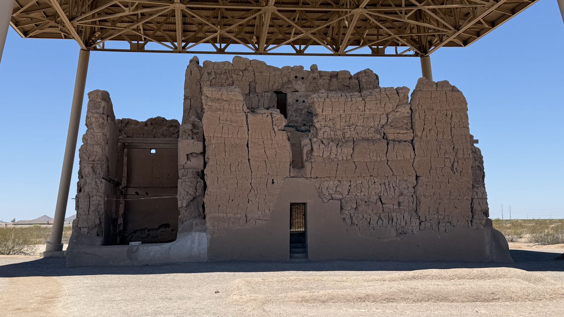 Desert adobe ruin beneath a large metal canopy; weathered mud-brick walls with a doorway and small windows, blue sky and barren landscape in the background.