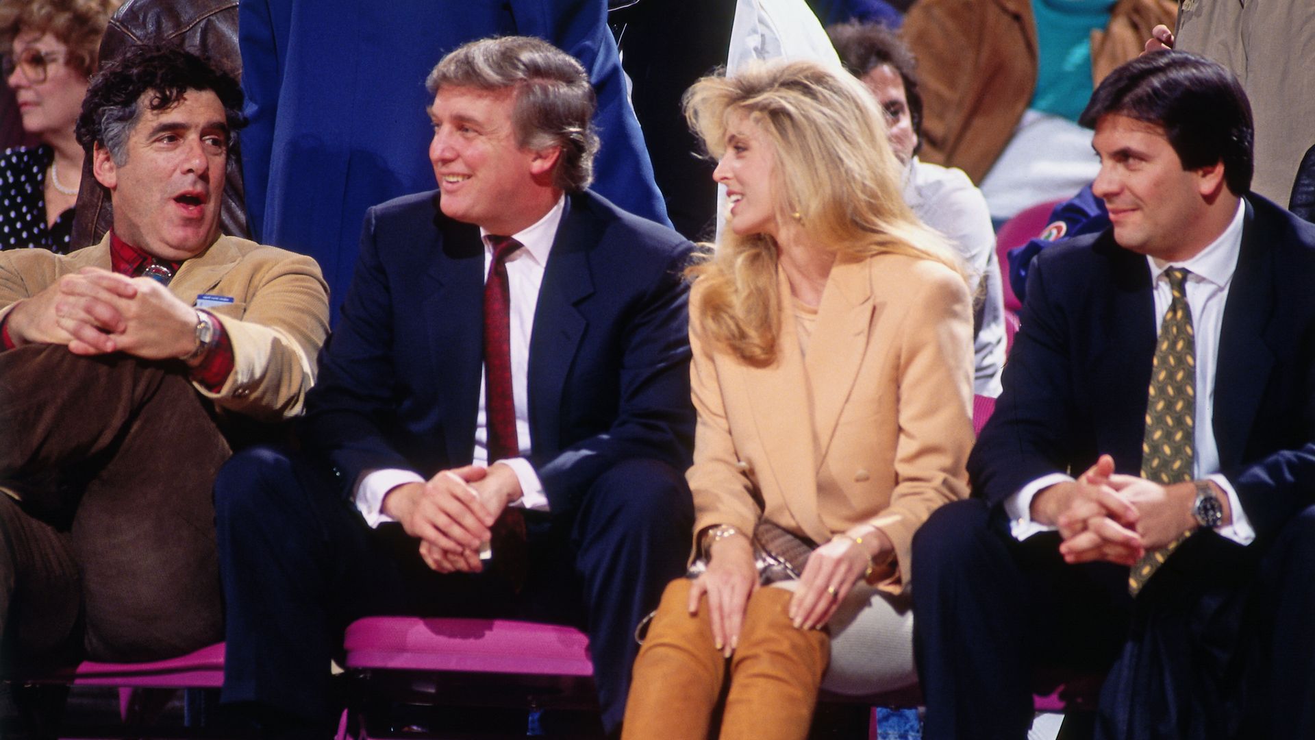 Four people sit in a row at an event, smiling. From left: person in a tan blazer, person in a navy suit, blonde person in a tan coat, and a person in a dark suit with a yellow tie; pink seats behind.