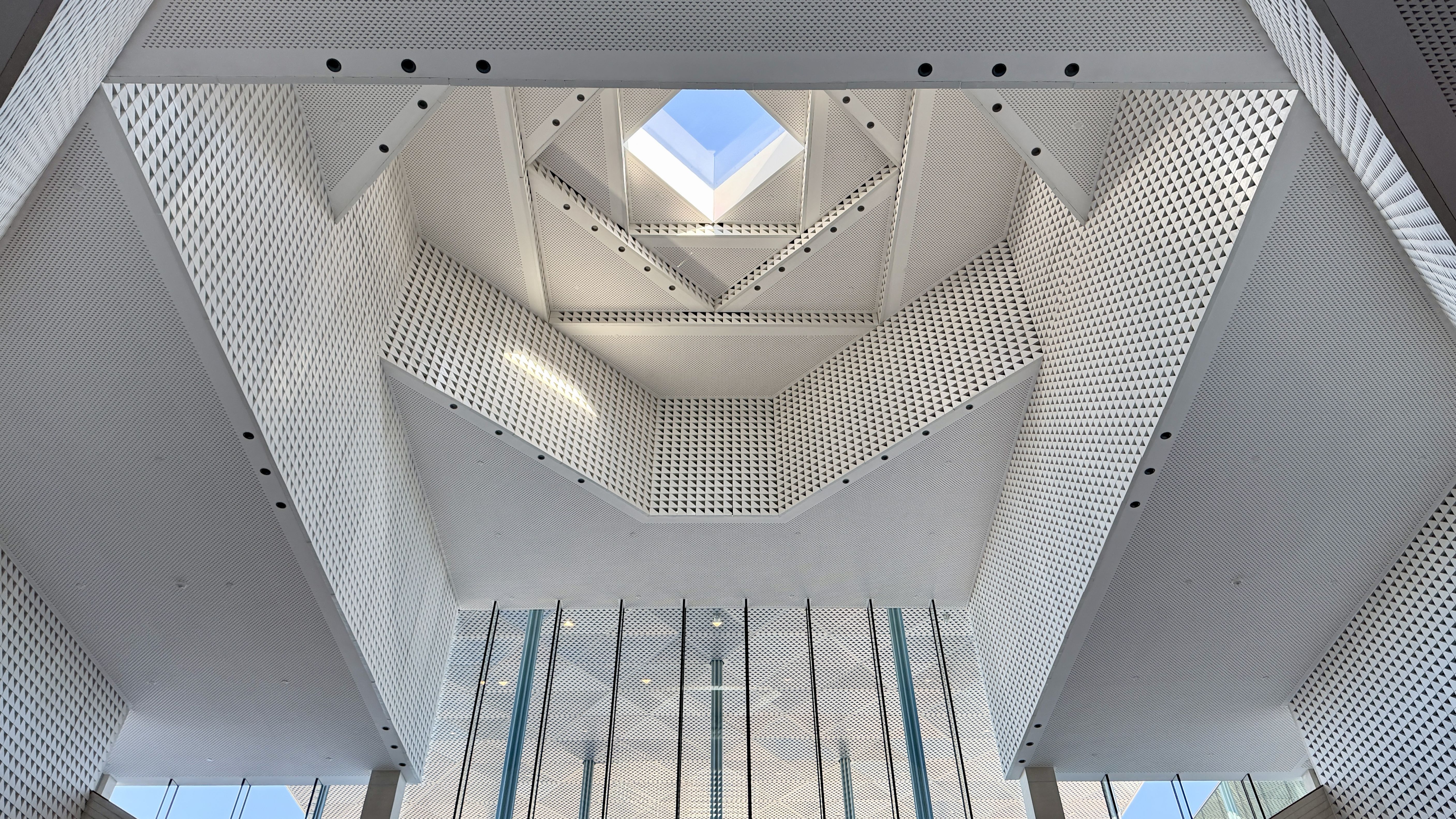 Interior ceiling view with geometric patterned white panels and a skylight opening showing blue sky, framed by modern architectural design with glass walls and columns.