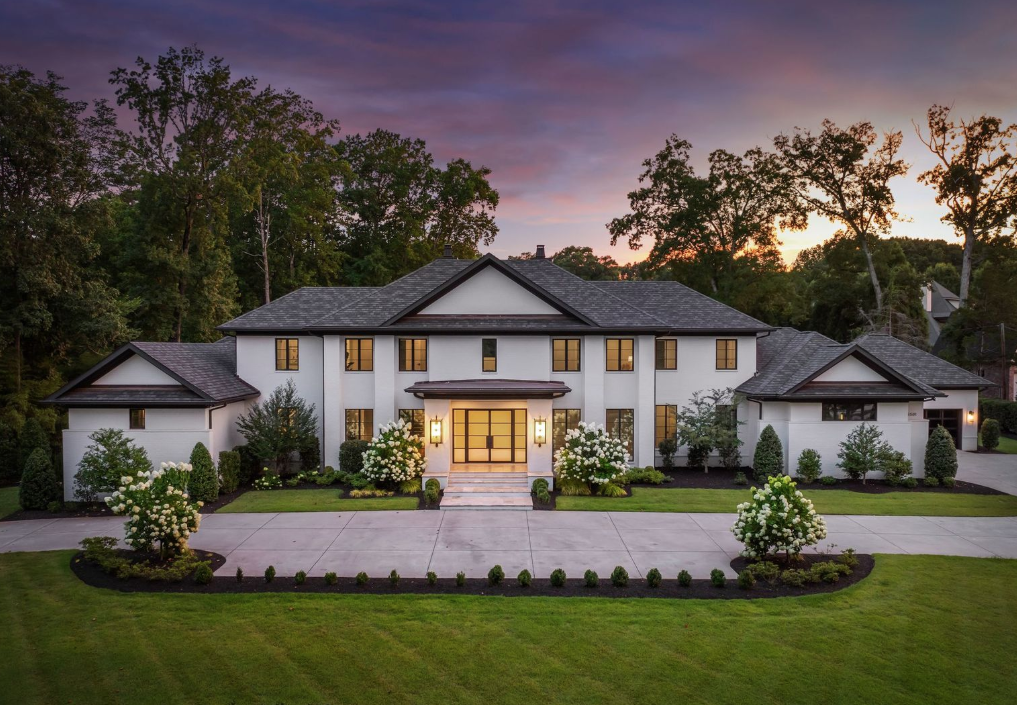 Large modern white two-story house with dark roof, symmetrical design, lit entrance, surrounded by green lawn, flowering bushes, and tall trees under a purple and orange sunset sky.