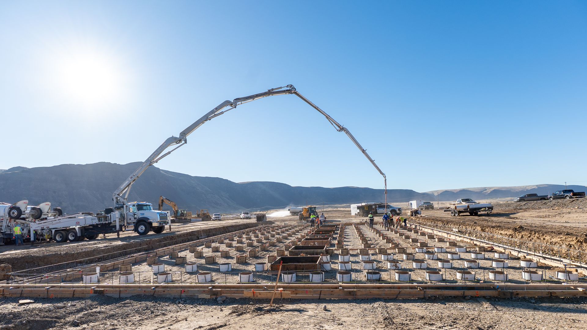 Construction site under clear blue sky with workers and machinery including a concrete pump truck pouring concrete into cylindrical forms arranged in a grid, surrounded by dirt and hills.