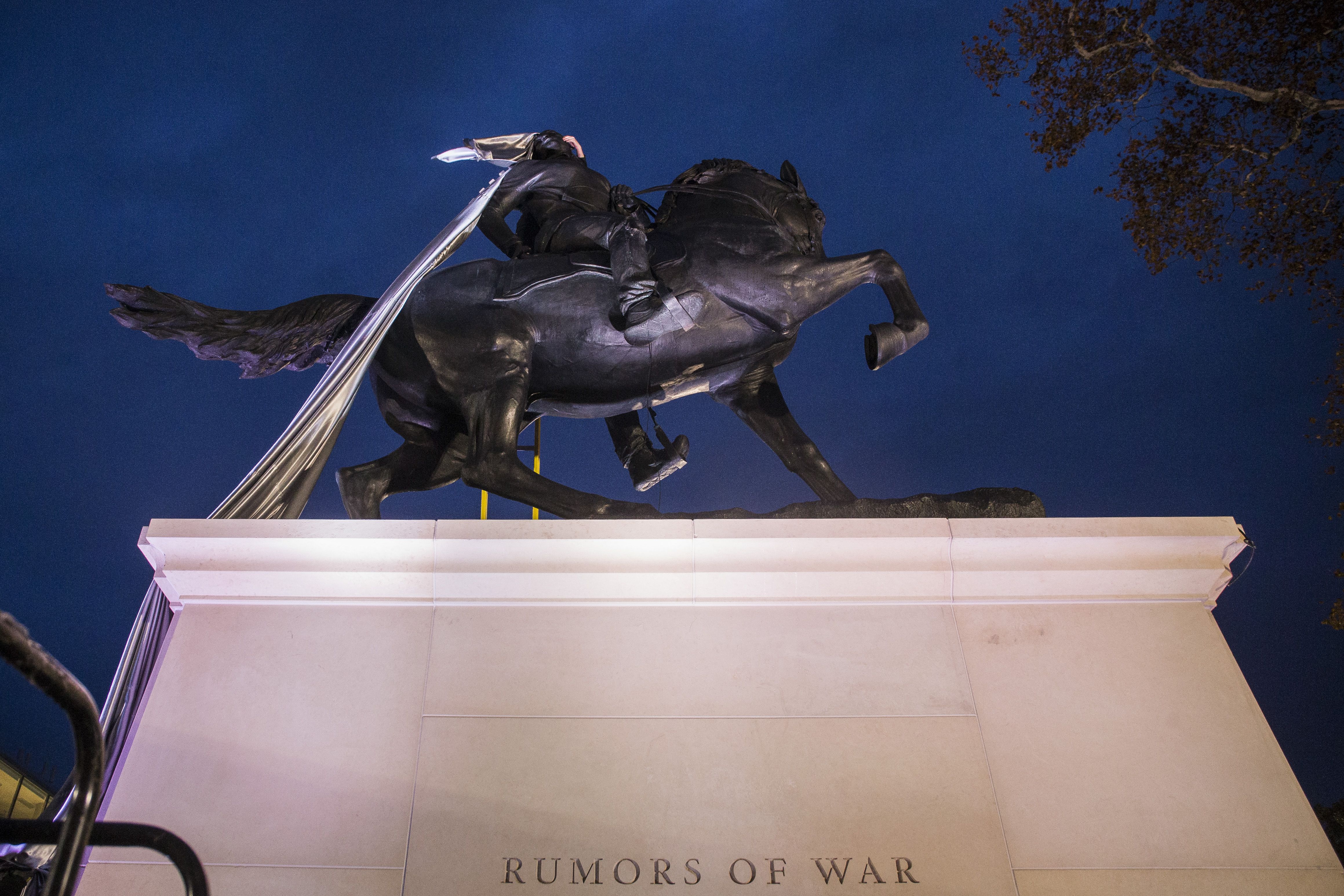 Bronze equestrian statue of a rider on a rearing horse atop a white pedestal, dark clothing and a long silver scarf streaming behind. Deep blue night sky; base reads "RUMORS OF WAR"