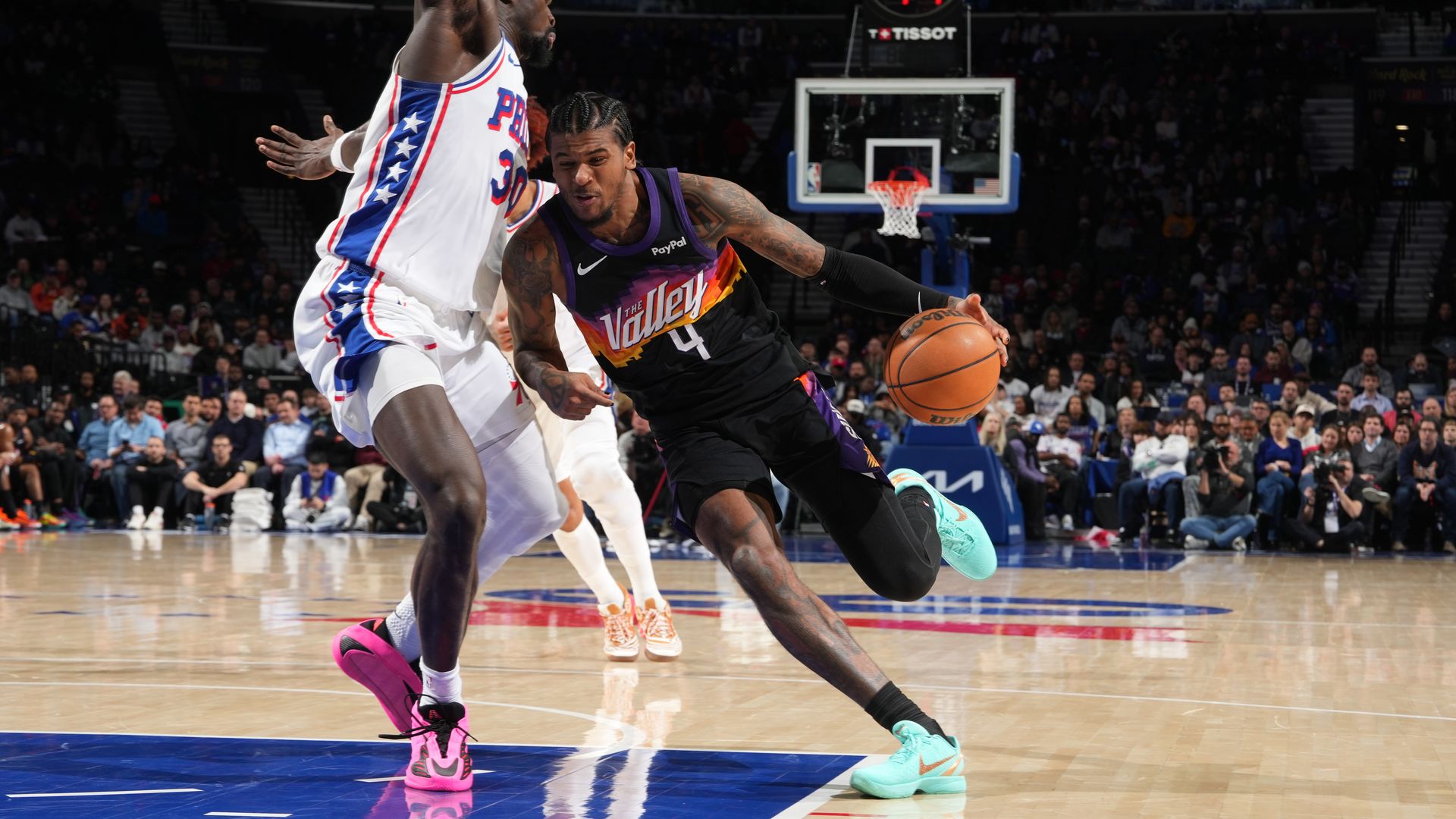 A basketball player in a black jersey with the word "Valley" over a red, orange and yellow design on the front over the number "4" drives with the ball against a defender in a white jersey with a thick blue stripe running down the side. 