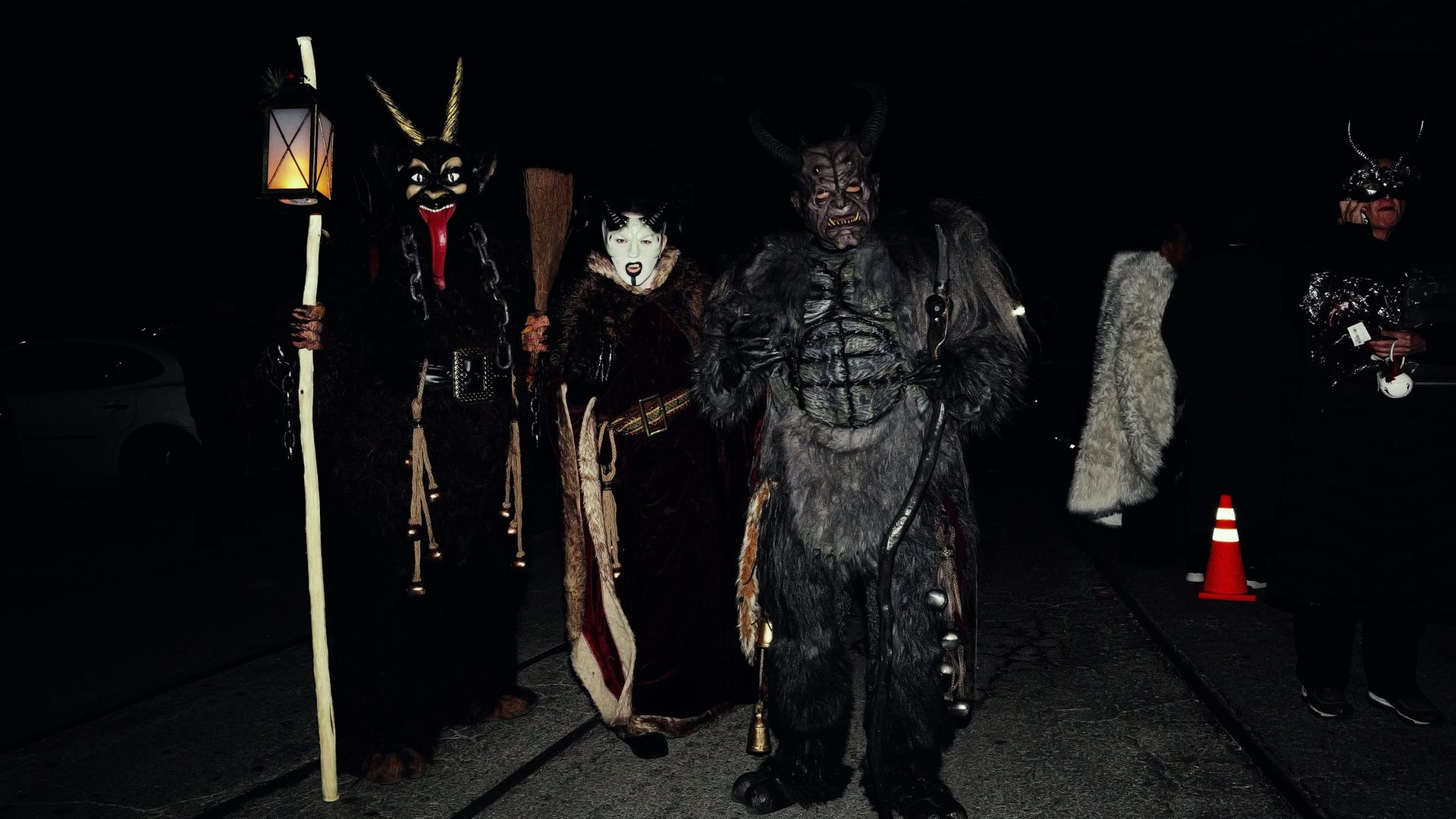 Three people in dark, furry demon costumes with horns and scary masks stand on a street at night, one holding a lantern and another a stick. Others in costumes are nearby at the inaugural 2024 Krampus Parade in San Antonio.
