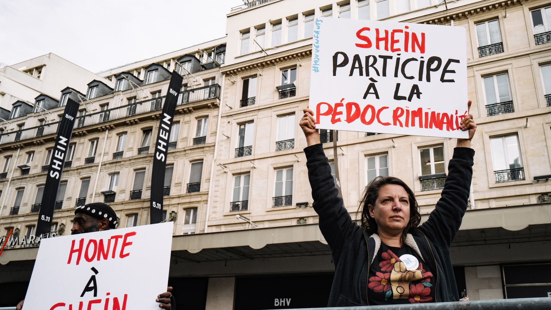 A woman holds a sign above her head that accuses Shein of participating in pedocriminality outside of a building with Shein signs.