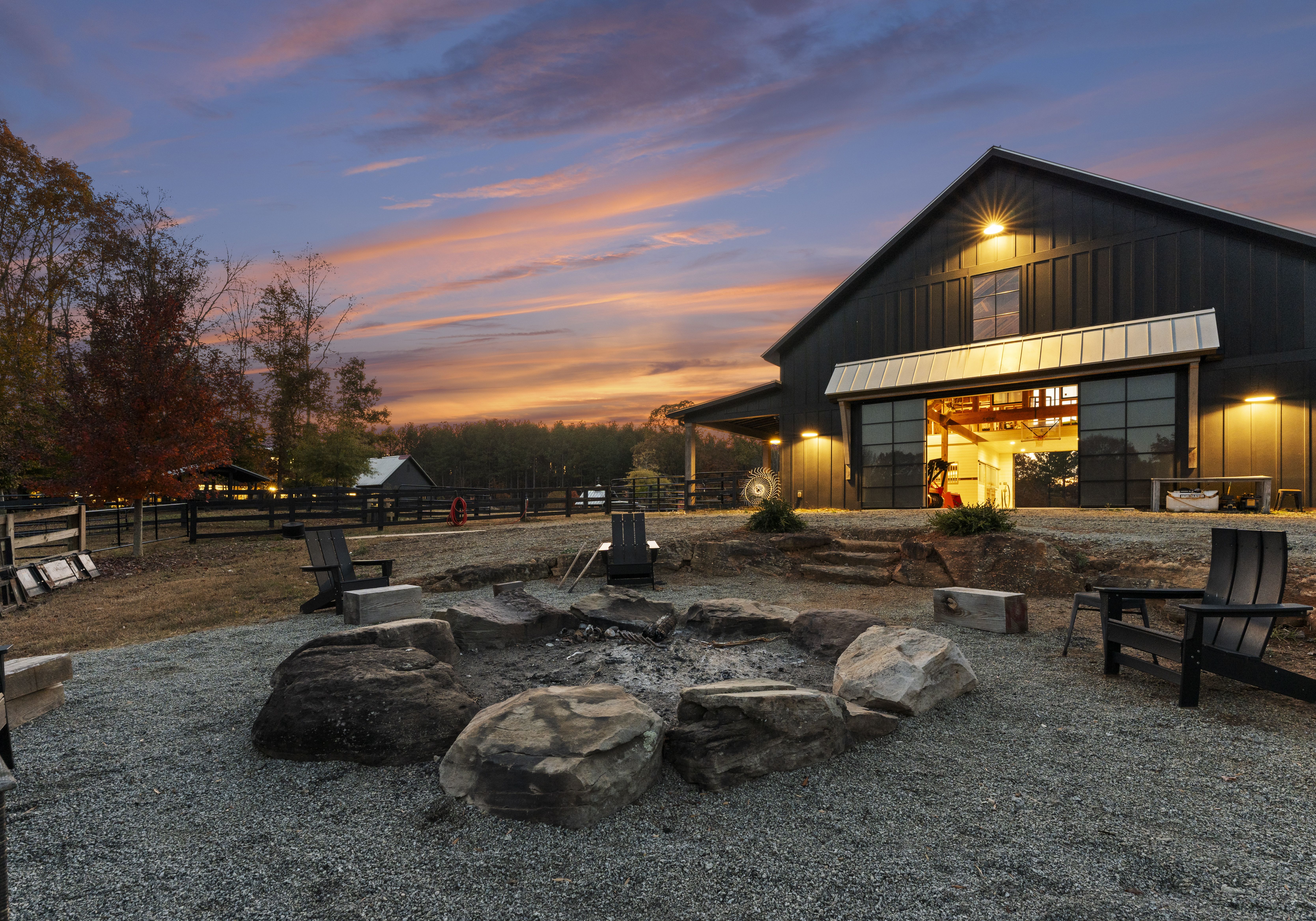 Sunset at a farm: a dark barn with warm interior lights and open doors, a rock fire pit ring in gravel, and Adirondack chairs, with autumn trees and a fence under an orange sky.