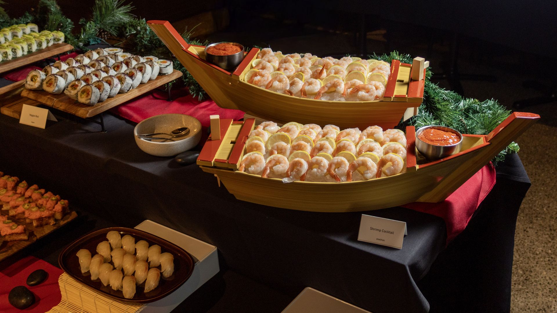 Assorted sushi and shrimp on decorative wooden boat platters with lemon wedges and dipping sauces, set on a dark table amid greenery in a dim buffet scene.