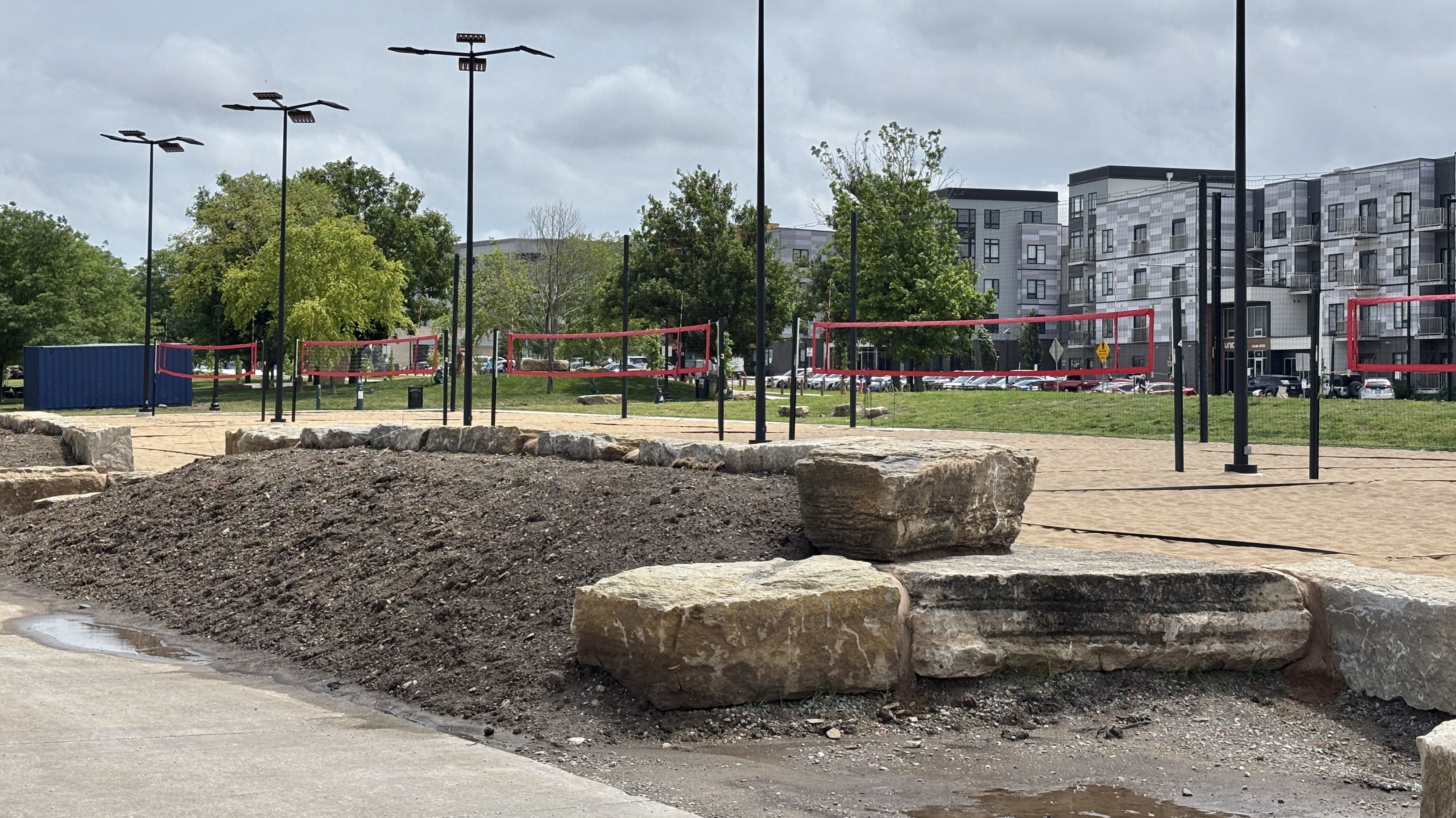 Public park with red volleyball nets, tall floodlights, green trees, and a row of modern gray apartment buildings in the background; rocky foreground and a cloudy sky.