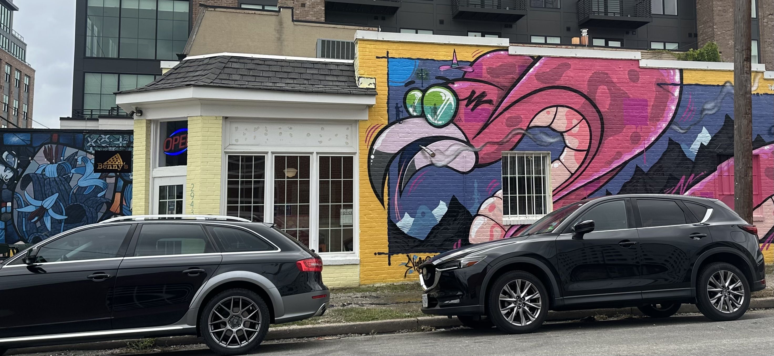 Colorful street mural on brick wall with yellow storefront, neon "OPEN" sign, and two gray SUVs parked in front; left panel blue abstract art, right panel pink and purple swirl.