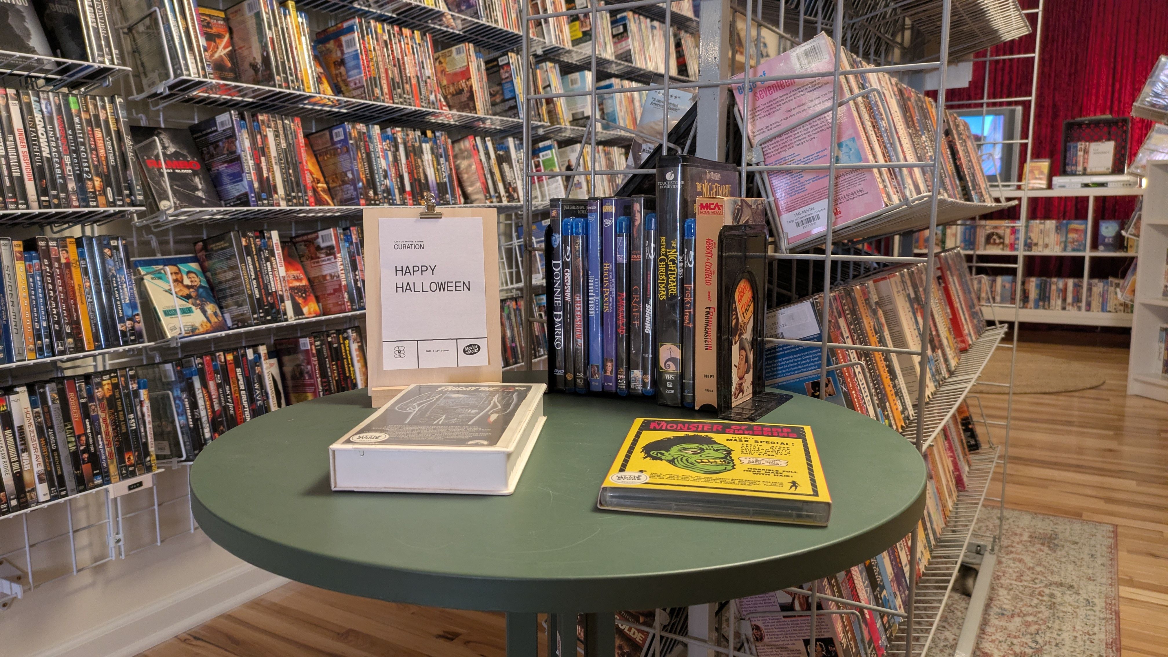 Inside a busy video store, metal shelves overflow with movie cases. A large round green table in the foreground holds a clipboard reading "HAPPY HALLOWEEN" as shelves and red curtains fill the background.