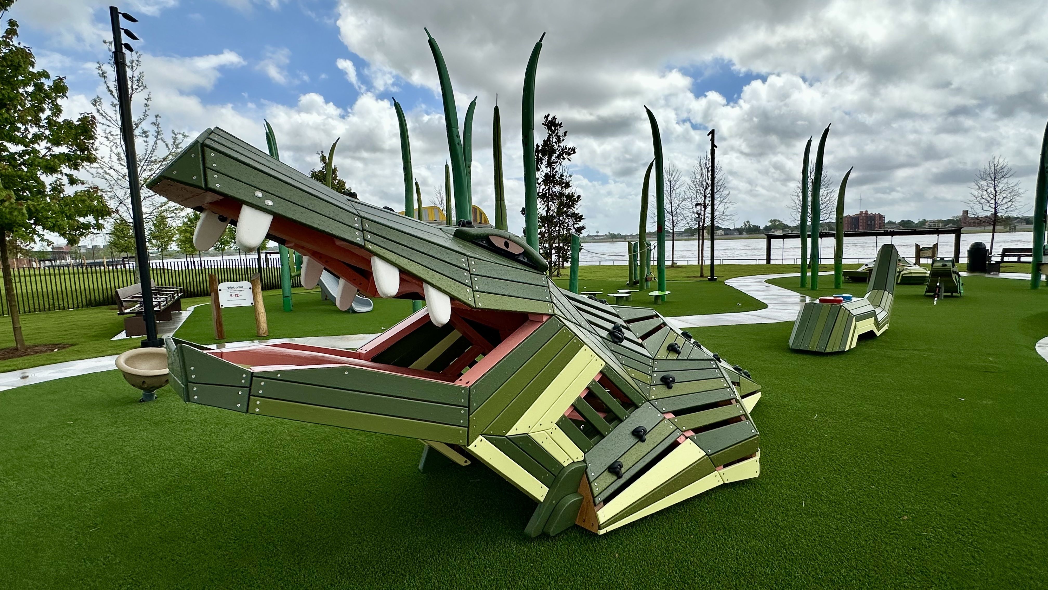 Green modular playground sculpture resembling a dragon with an open mouth and white teeth sits on artificial grass by a river, with a curved path, tall green posts, benches nearby.