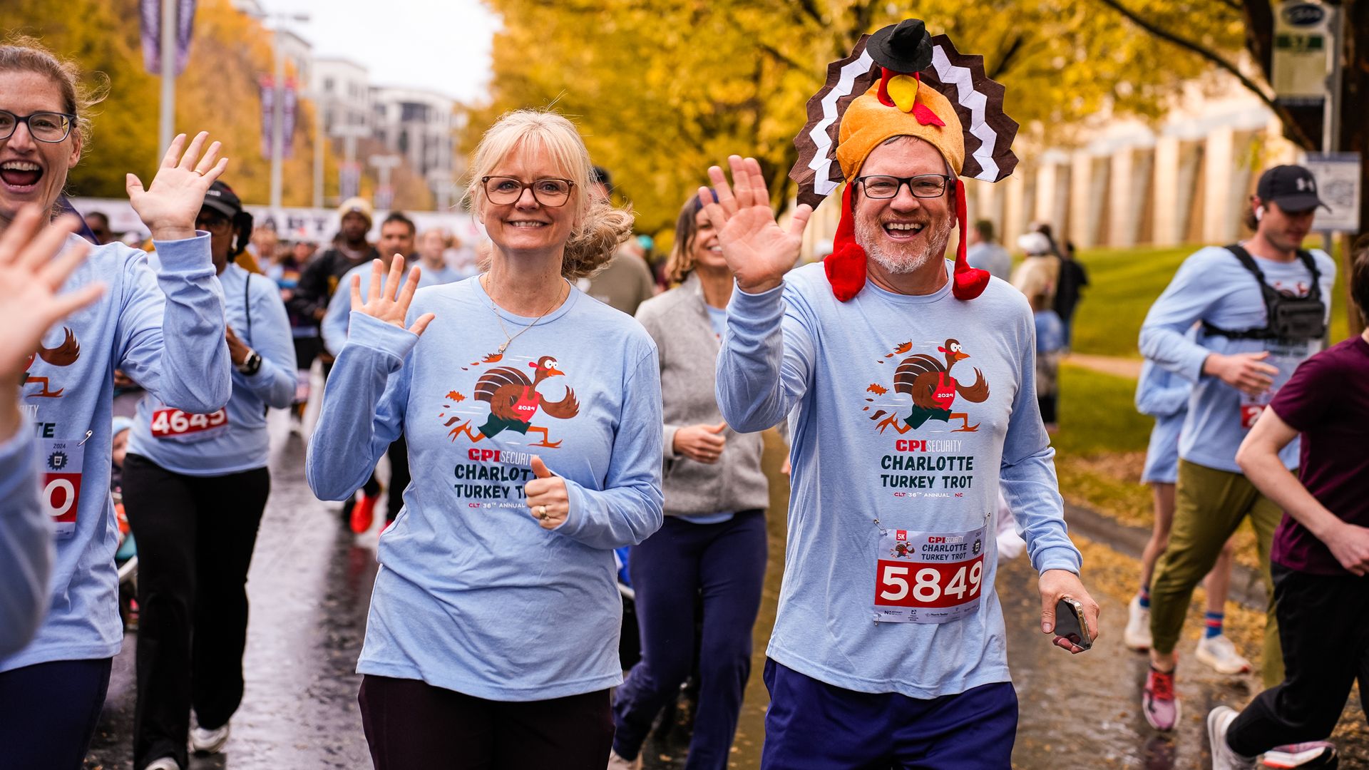 People running a fall race with yellow autumn trees in the background. Two front runners smile and wave, wearing light blue shirts with a turkey graphic; one man wears a turkey hat.
