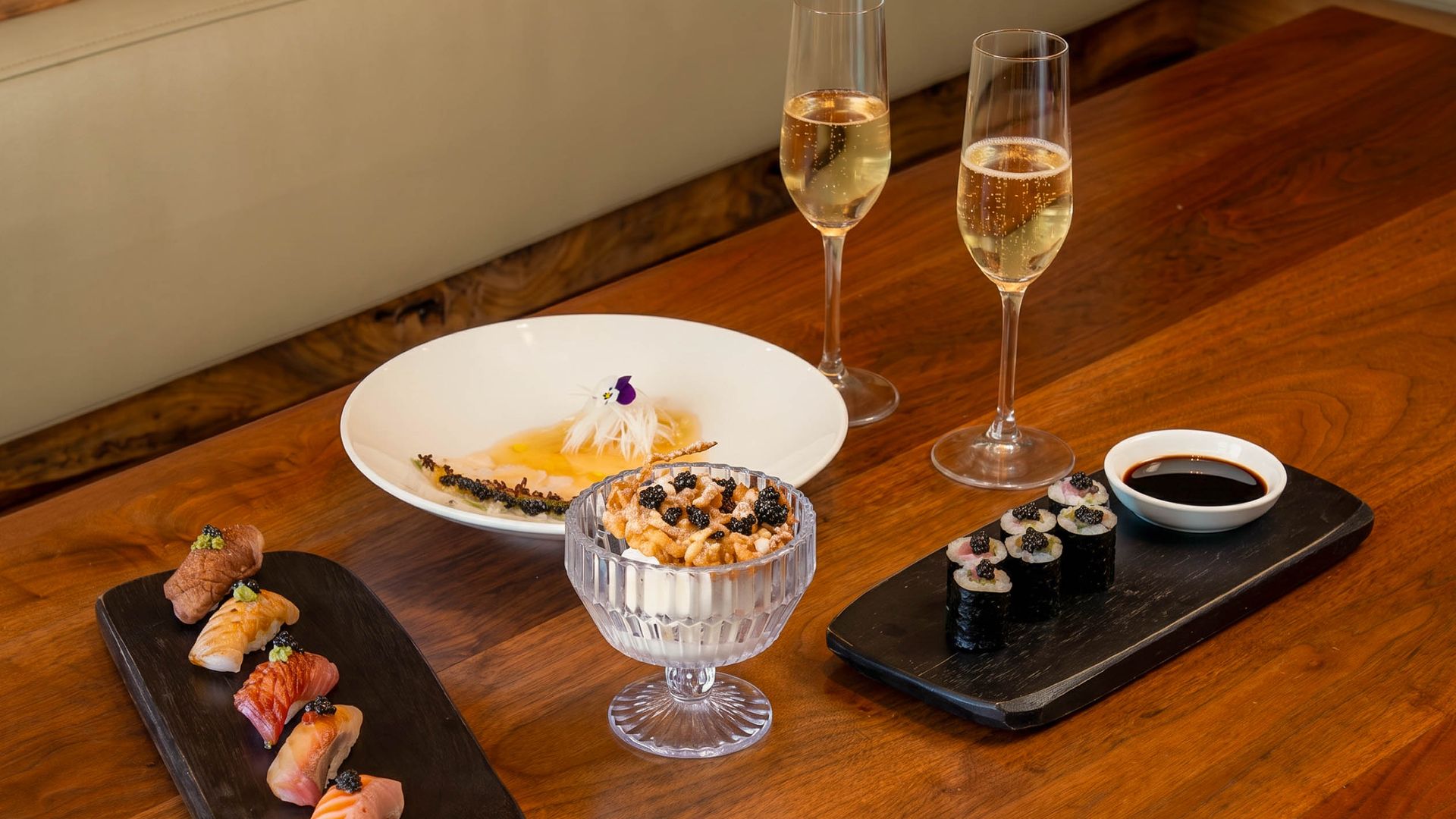 Two champagne flutes and a white dessert plate sit on a wooden table, with sushi rolls, pastries, and a small soy sauce cup in a cozy dining setting.