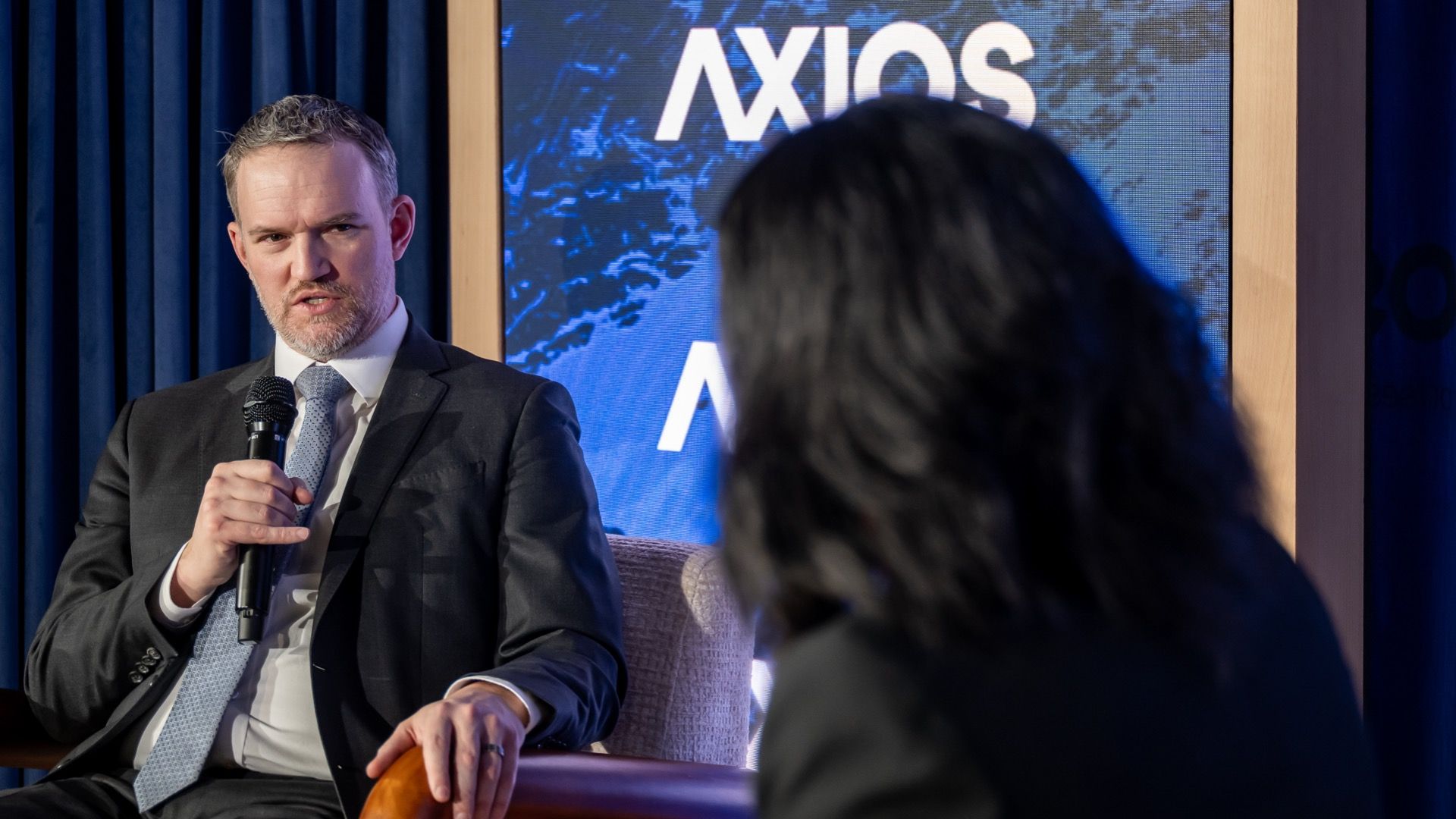 A man in a dark suit and light blue tie speaks into a microphone during an Axios event, with a seated woman with dark hair in the foreground and an Axios logo backdrop in blue and white.